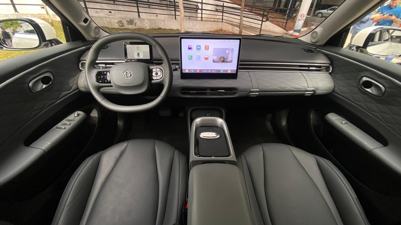 Interior de um carro moderno, com bancos de couro cinza escuro, volante multifuncional e duas telas digitais no painel, uma atrás do volante e outra central maior exibindo ícones de aplicativos. O console central tem acabamento metálico e preto, e as portas possuem detalhes acolchoados com padrão de losangos. Ao fundo, fora do carro, vê-se um homem de camisa azul e parte de uma rampa