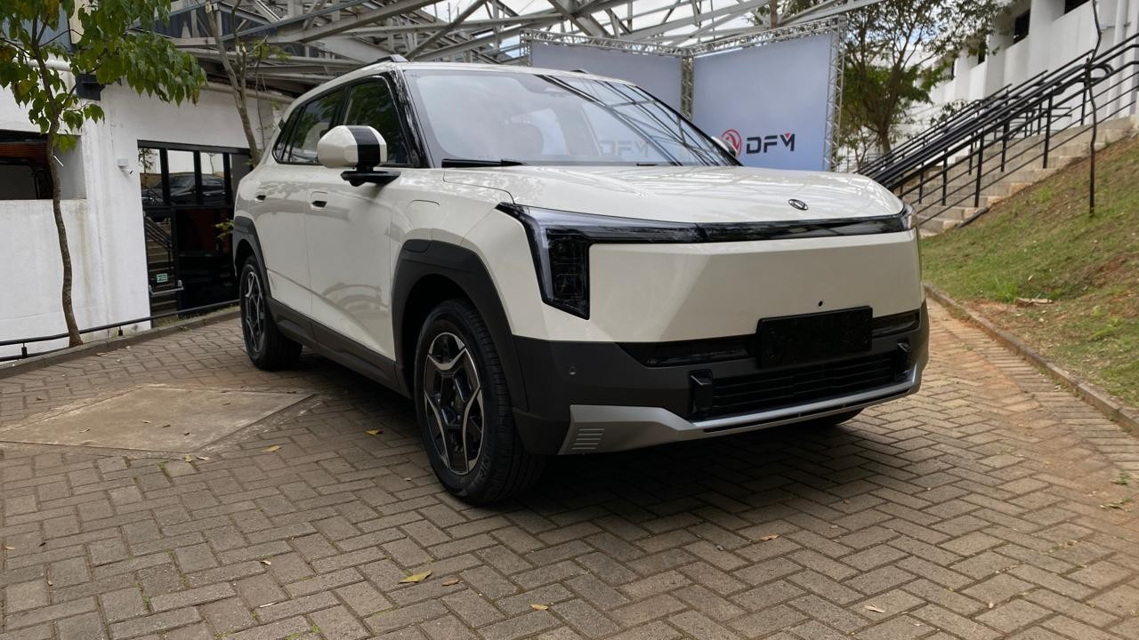 SUV elétrico branco com detalhes pretos e rodas escuras, estacionado em piso de tijolos, com uma estrutura metálica e um banner branco com o logo DFY ao fundo