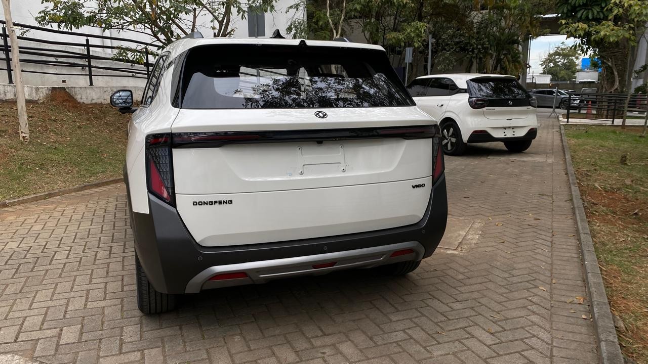 Traseira de um carro SUV branco da marca Dongfeng, com detalhes em preto e cinza, estacionado em um piso de blocos de concreto. Ao fundo, outro carro branco menor e árvores.
