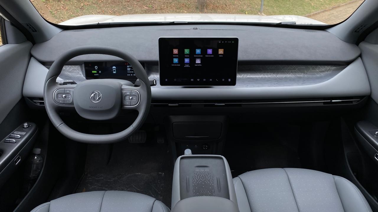Interior de um carro moderno com volante cinza, painel digital e uma grande tela multimídia central exibindo ícones de aplicativos. O console central possui um carregador por indução e os bancos são cinza claro.