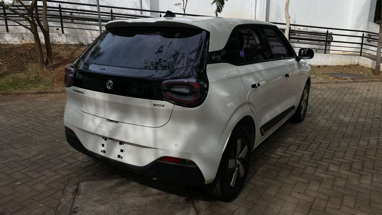 Carro elétrico branco com teto e detalhes pretos, visto de traseira e lateral, estacionado em piso de blocos de concreto, com vegetação e muro ao fundo