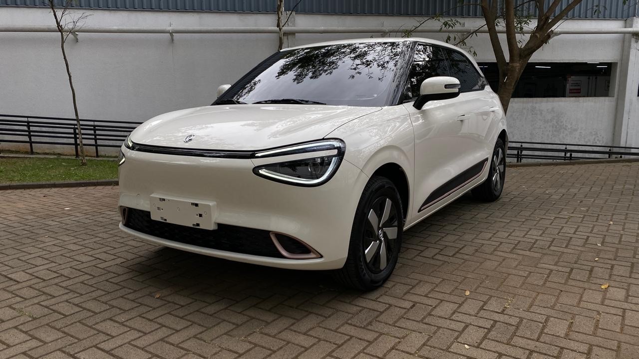 Carro elétrico branco, modelo SUV compacto, estacionado em piso de blocos de concreto. O veículo tem faróis de LED em formato de C e rodas com detalhes em preto e prata. Ao fundo, uma parede clara, vegetação e uma rampa de acesso.