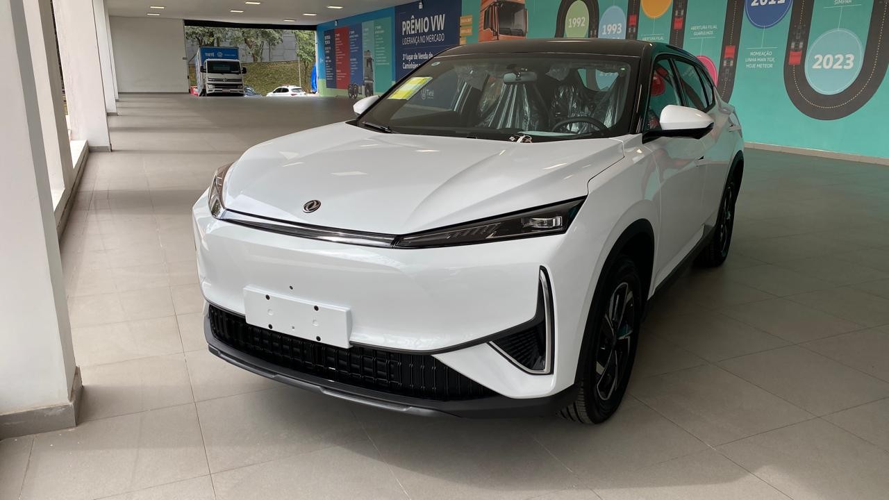 Carro branco moderno, com teto preto e rodas escuras, exibido em um showroom com piso claro e paredes decoradas com gráficos de linha do tempo e o texto PRÊMIO VW