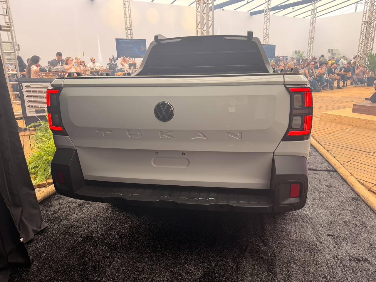 Traseira de uma picape branca com o nome TUKAN em relevo e o logo da Volkswagen, lanternas verticais acesas em vermelho, para-choque preto. Pessoas e estruturas metálicas desfocadas ao fundo