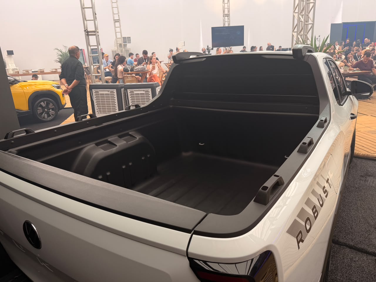 Traseira de uma picape branca com caçamba vazia e aberta, exibindo o revestimento preto. Na lateral, a palavra ROBUST em cinza. Ao fundo, pessoas em um evento, um homem de camisa preta e uma picape amarela