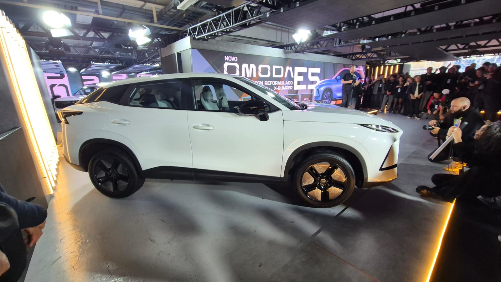 SUV branco Omoda E5 em destaque num palco iluminado, com o letreiro NOVO OMODA E5 ao fundo. Pessoas e fotógrafos registram o evento.