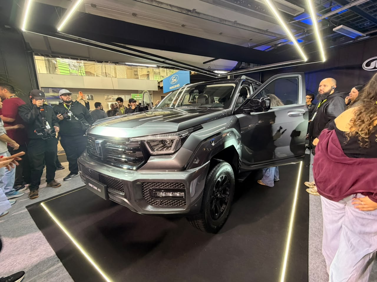 Um SUV cinza escuro com a porta do motorista aberta, exibido em um estande com luzes de LED no chão e teto. Pessoas observam o veículo, algumas tirando fotos. Ao fundo, um banner azul com a marca Ford