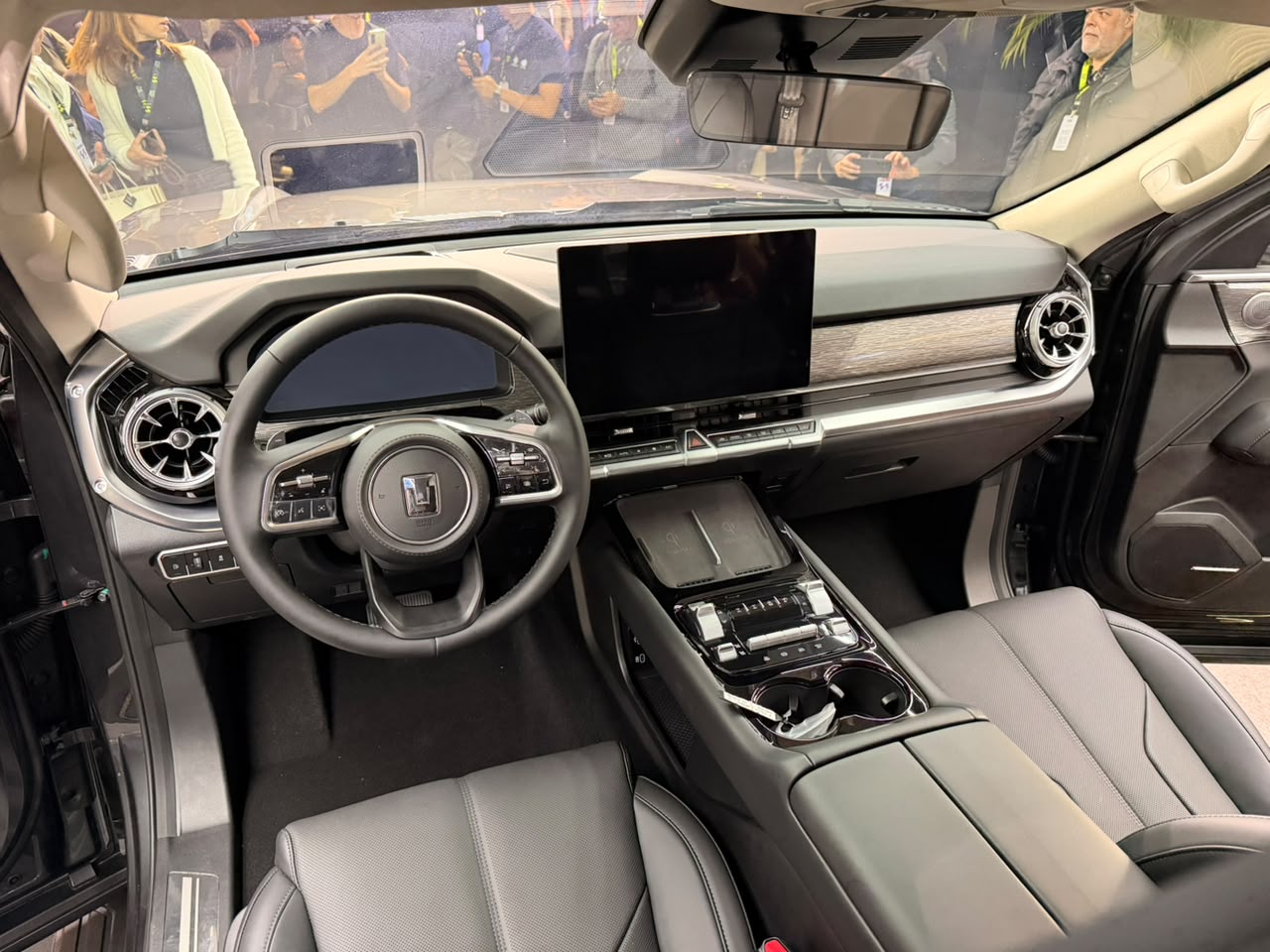Interior de um carro moderno com volante multifuncional, tela central grande e painel com acabamento em madeira clara. Os bancos são de couro cinza escuro e o console central possui porta-copos e carregador por indução. Pessoas borradas aparecem ao fundo, refletidas no para-brisa