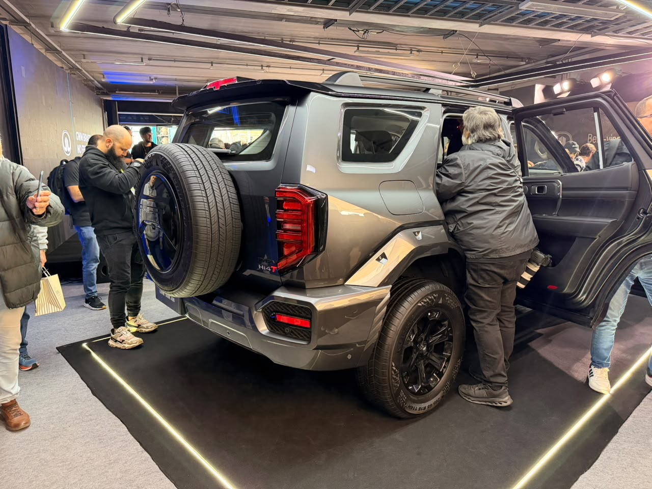 Pessoas observam um SUV cinza-chumbo com pneu estepe na traseira, em um estande de exposição. Um homem de jaqueta preta entra na porta traseira aberta do veículo, enquanto outros visitantes estão próximos, alguns com sacolas. O carro está sobre um tapete preto com iluminação de LED amarela, em um ambiente interno com luzes no teto