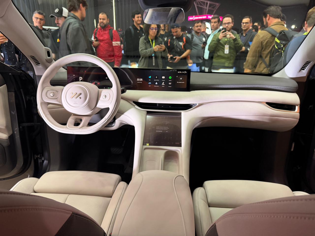 Interior de carro moderno com volante e painel brancos, bancos de couro bege. Uma tela horizontal exibe ícones de aplicativos. Ao fundo, um grupo de pessoas observa o veículo, algumas tirando fotos