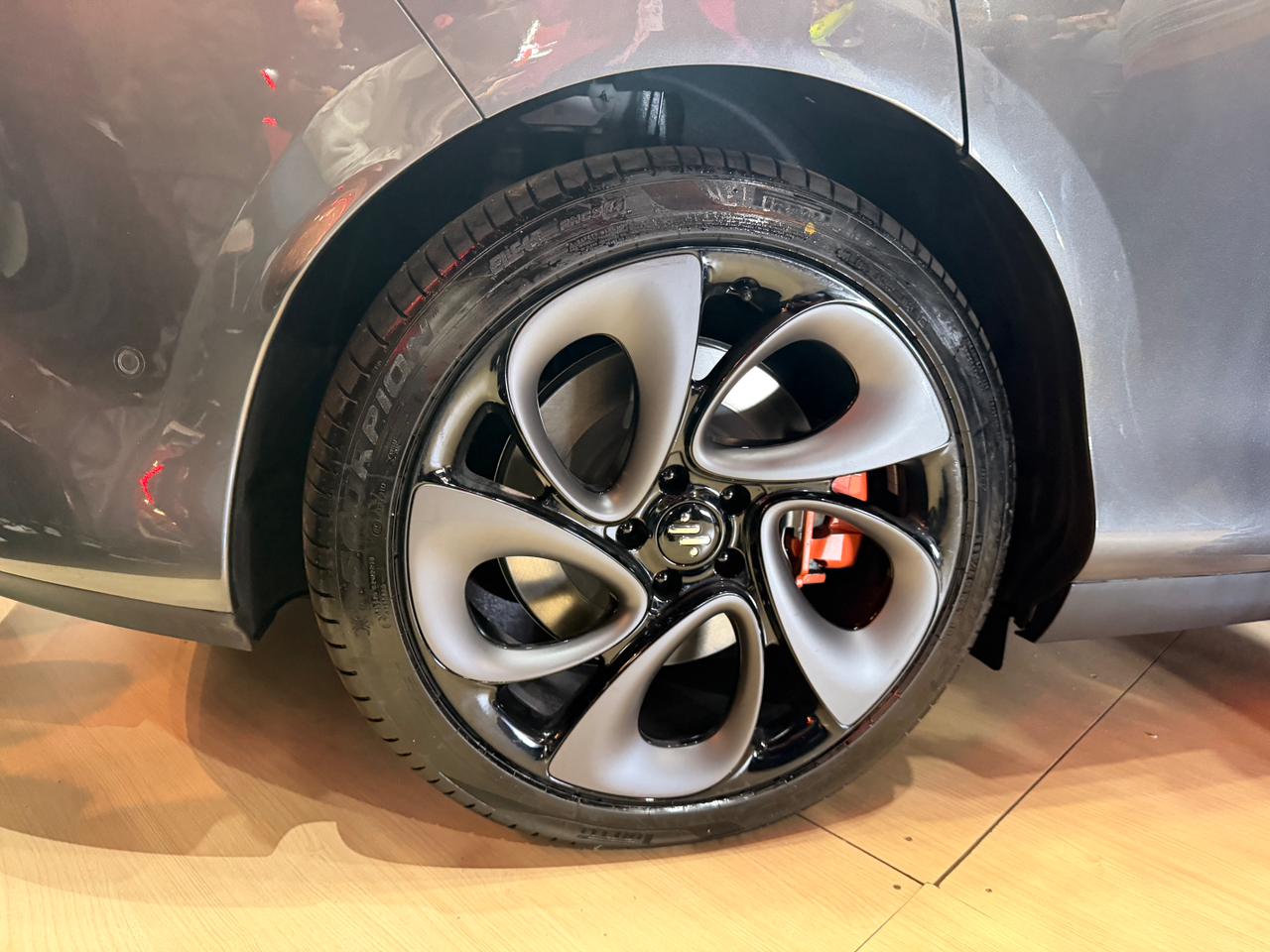 Roda de carro com design moderno, aro preto brilhante e cinco raios cinza-claro em formato de bumerangue, com pinça de freio laranja visível ao fundo. O pneu preto tem a marca Pirelli e o carro é cinza-chumbo, refletindo luzes vermelhas. O veículo está sobre um piso de madeira clara.