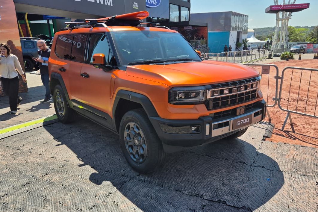 SUV laranja fosco com prancha de surf no teto, estacionado em área externa com piso cinza e pessoas ao fundo. O veículo tem grade frontal preta e cromada, faróis retangulares e para-choques robustos. À direita, uma cerca metálica e terreno de terra batida.