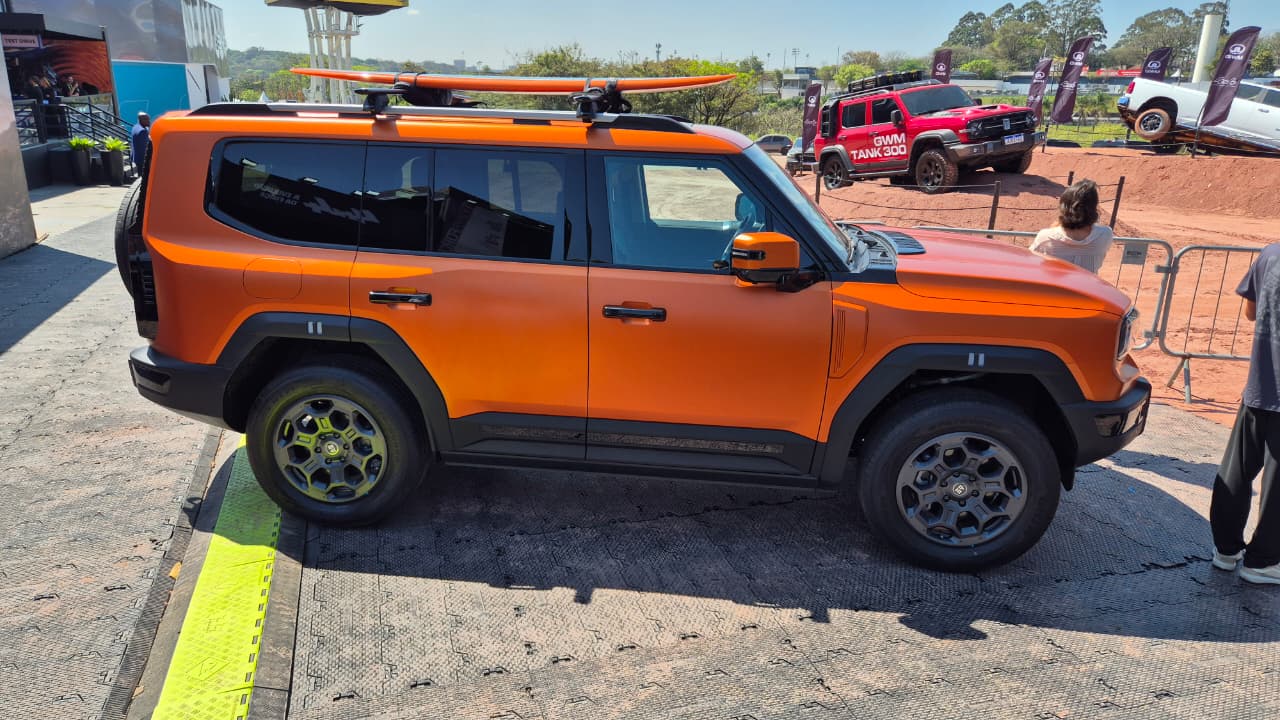 SUV laranja fosco com prancha de surf no teto, estacionado em área externa. Ao fundo, outros veículos off-road, um vermelho e um branco, em terreno acidentado, e pessoas observando. O dia está ensolarado
