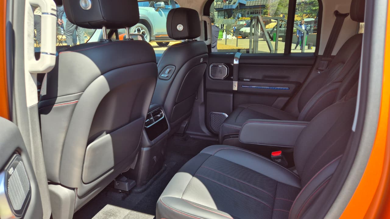 Interior de um carro laranja, mostrando os bancos traseiros pretos com detalhes em cinza e costura vermelha. Há um apoio de braço central rebatido, saídas de ar e controles no console traseiro. A janela exibe um cenário externo com pessoas e outros veículos