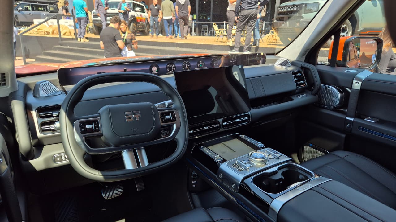 Interior de um carro moderno, com volante escuro e detalhes prateados, painel digital amplo e console central com tela sensível ao toque e botões giratórios. Ao fundo, pessoas e outros veículos em um evento ao ar livre