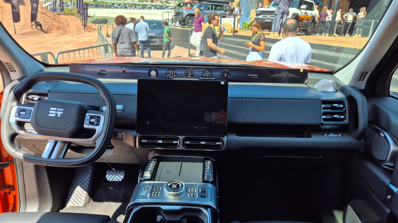 Interior de um carro moderno, com volante multifuncional e tela central grande. Ao fundo, pessoas em um evento ao ar livre com carros e rampas de terra