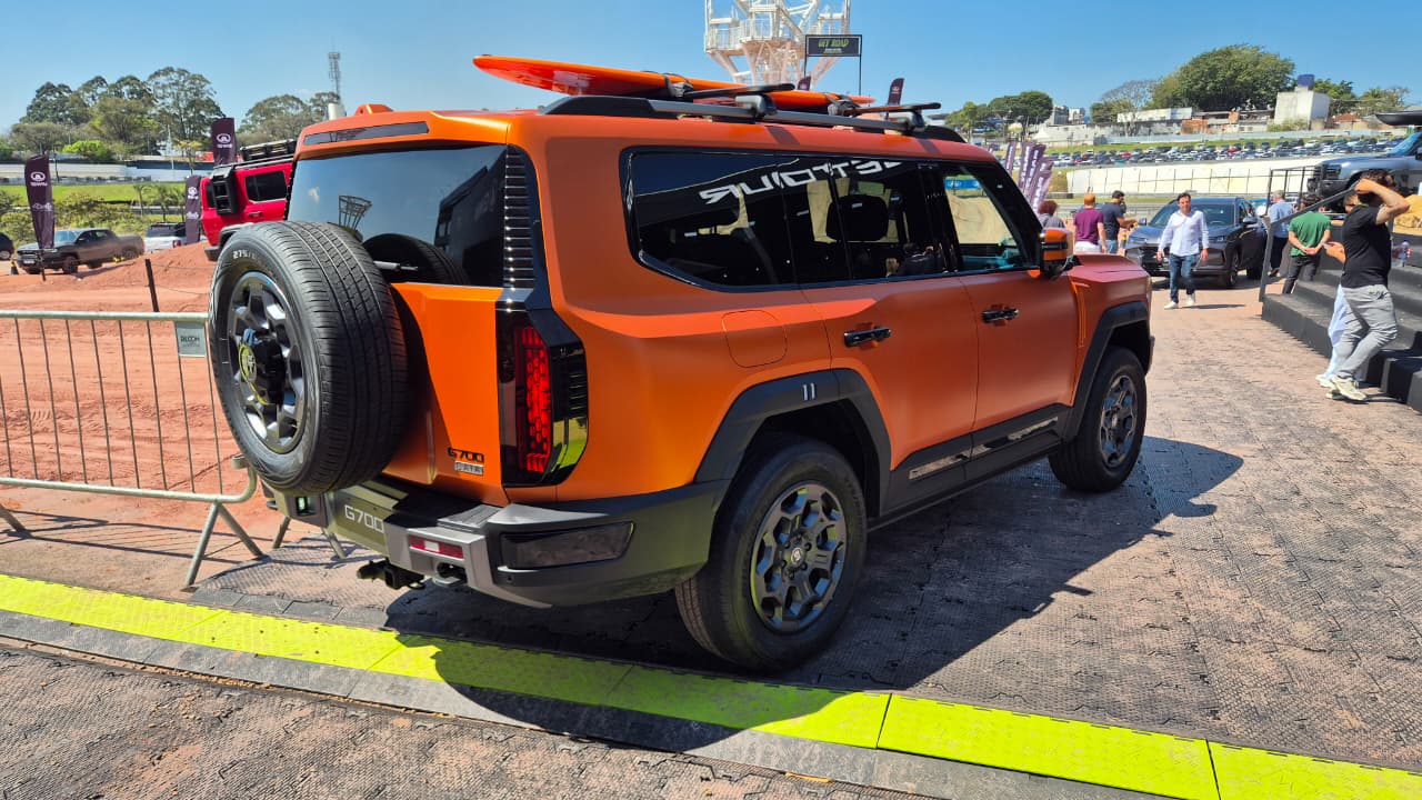 SUV laranja fosco com estepe na traseira e prancha de surfe no teto, estacionado em área externa com pessoas e outros veículos ao fundo, sob céu azul