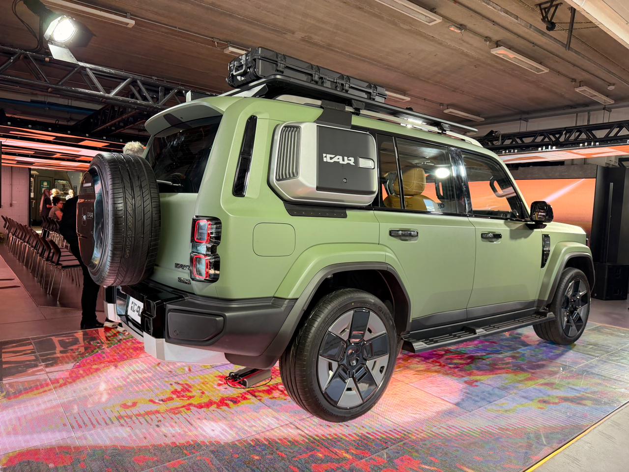 SUV verde-oliva com estepe na traseira, bagageiro de teto preto e um compartimento lateral cinza com a palavra TAUR em destaque, exibido em um evento com piso colorido e pessoas ao fundo