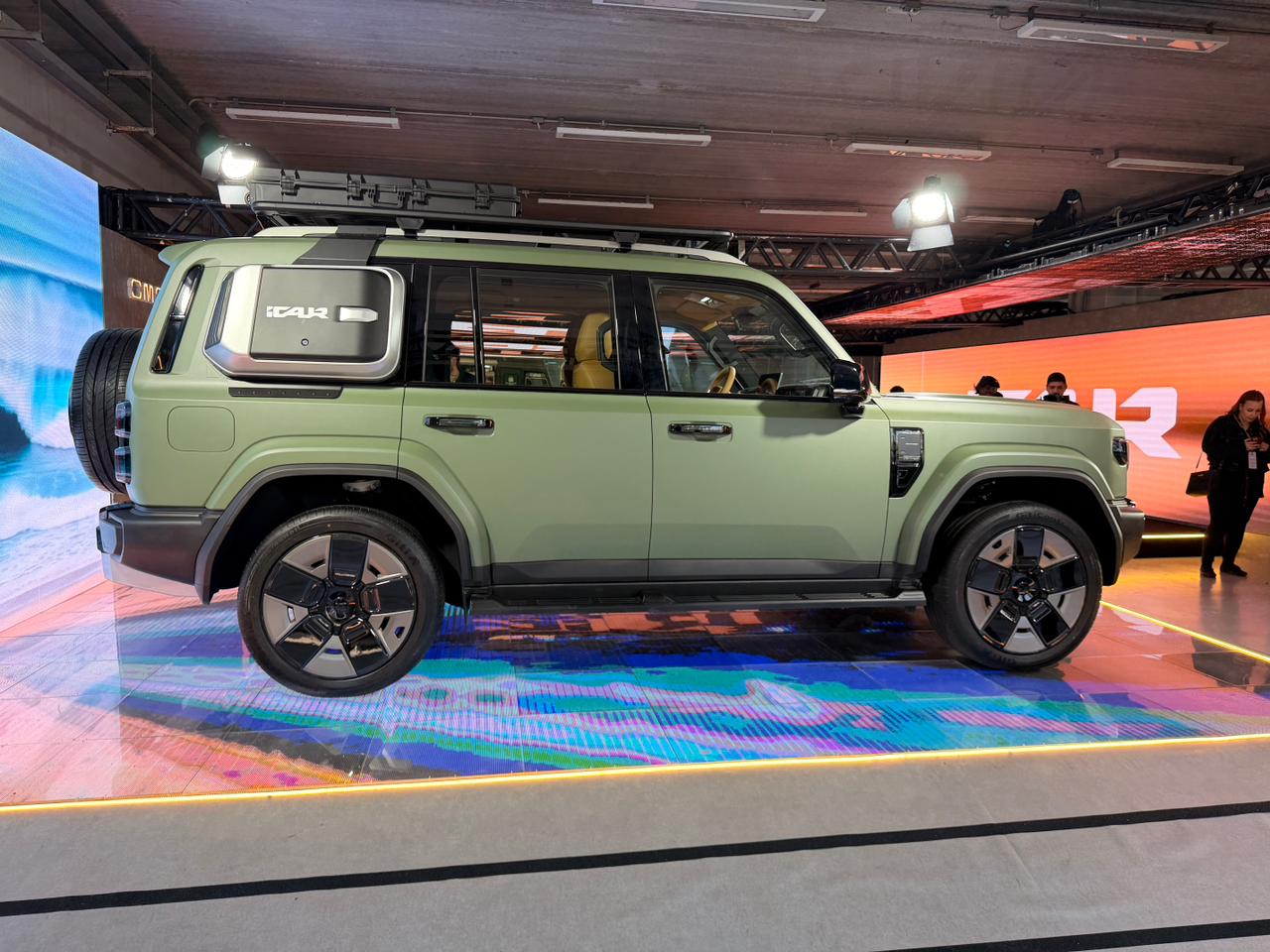 SUV verde-oliva com estepe na traseira, rack de teto e rodas pretas e prateadas, exibido em um palco iluminado com luzes coloridas. Pessoas observam ao fundo