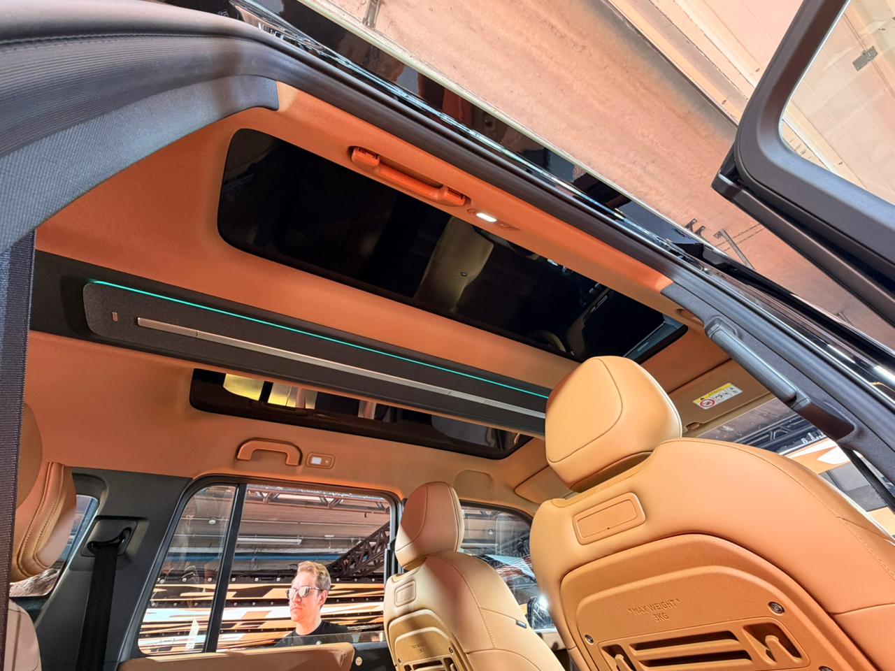 Interior de um carro com teto solar panorâmico e estofamento bege. Um homem de óculos e camiseta preta é visível através da janela traseira, refletindo o ambiente externo. Detalhes de iluminação ambiente em azul e laranja no teto e alças de segurança são visíveis