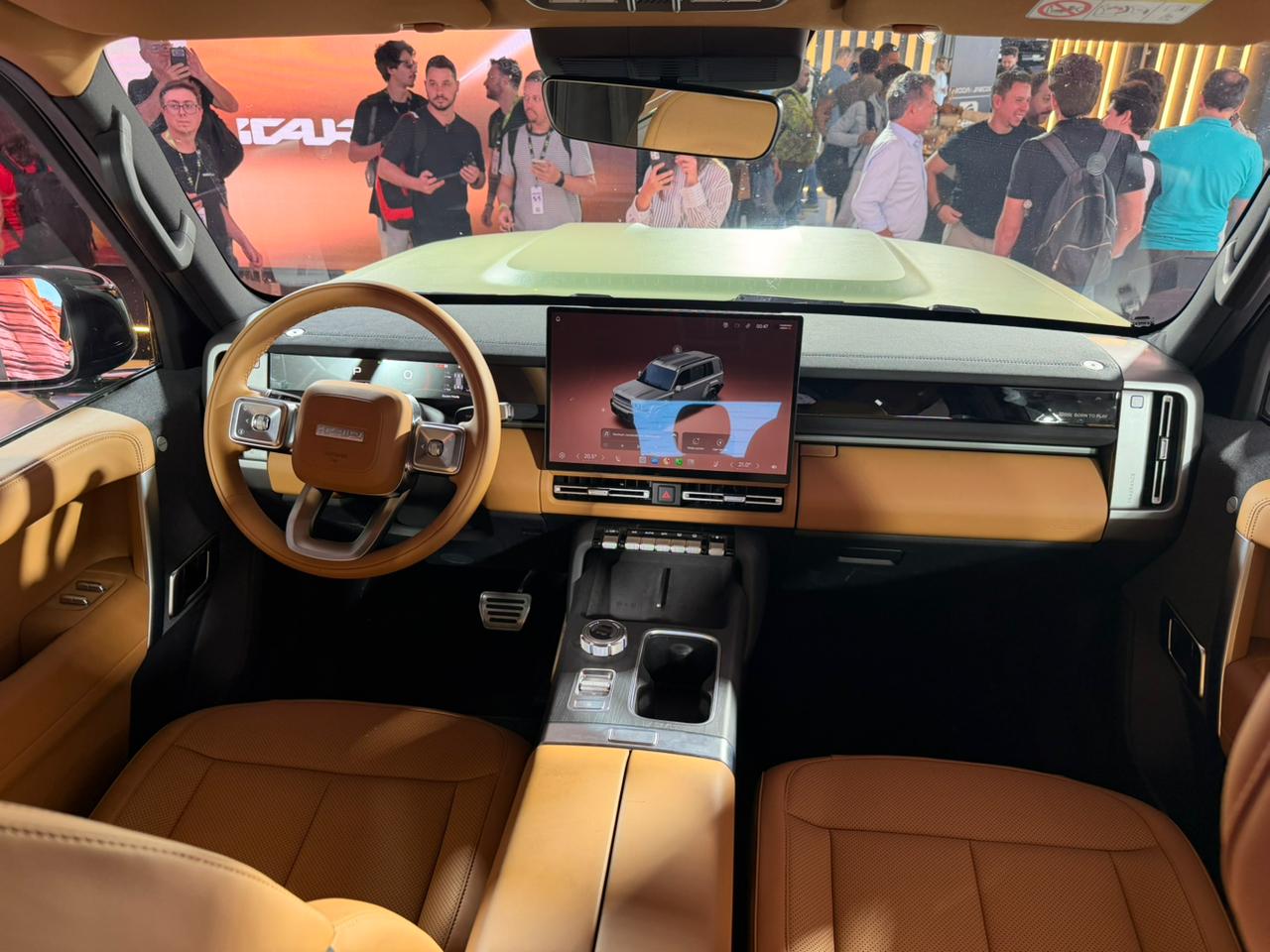 Interior de um carro moderno com volante marrom e painel em tons de bege e preto. Uma tela central exibe um SUV cinza. Pessoas em segundo plano observam o veículo.