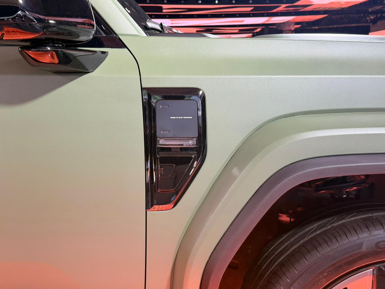 Lateral de um carro verde fosco, com retrovisor preto brilhante e um detalhe retangular escuro no para-lama dianteiro, acima da roda preta. O fundo é um borrão laranja e preto