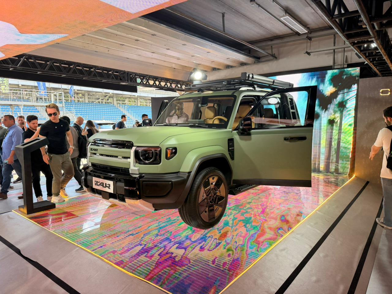 Um SUV verde-oliva com portas abertas e teto solar está em destaque sobre um tapete colorido, com pessoas ao redor em um evento. Ao fundo, uma tela exibe palmeiras