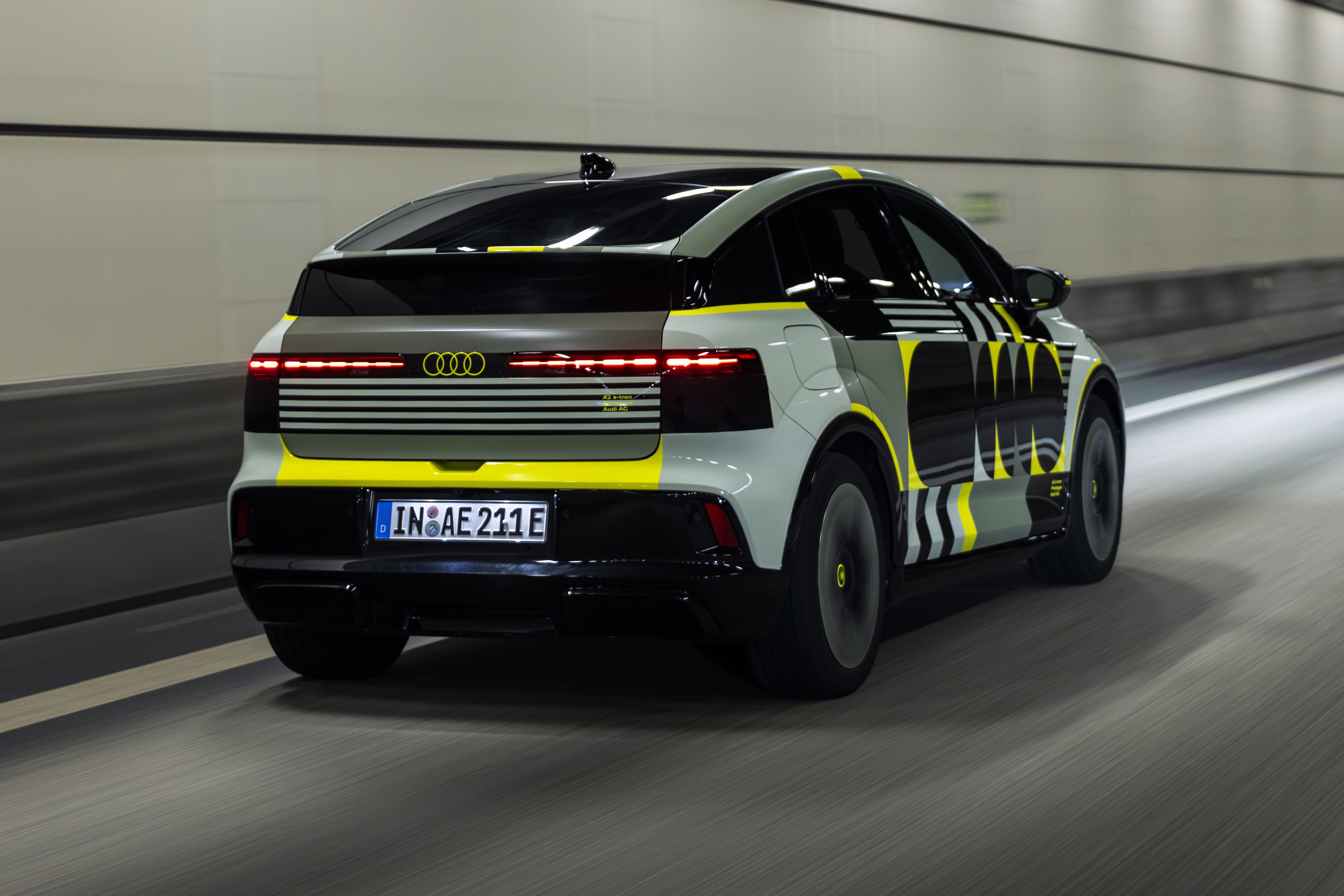 Carro Audi camuflado em cinza, preto e amarelo neon, visto por trás e lateralmente, em movimento rápido dentro de um túnel iluminado