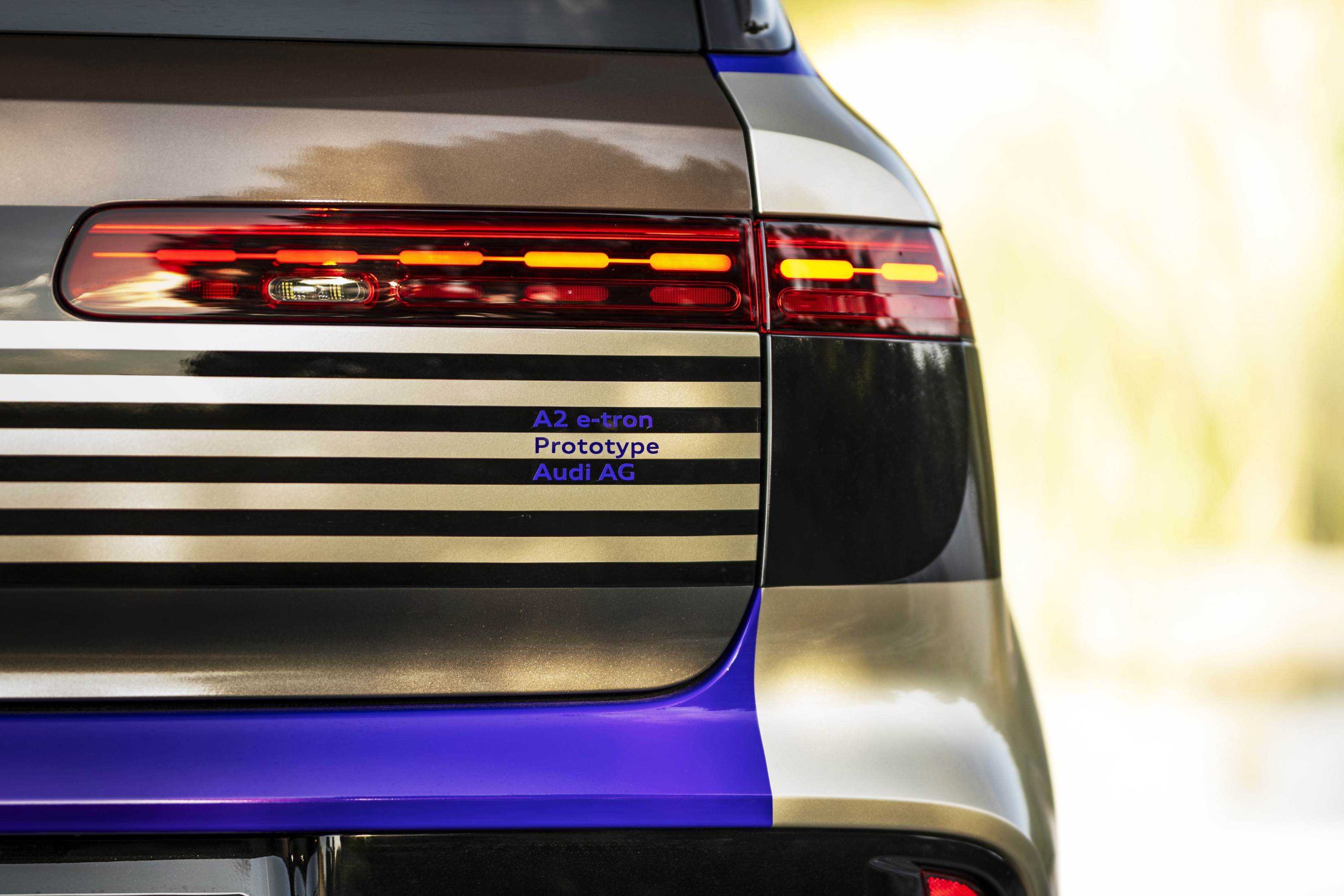 Traseira de um carro protótipo Audi A2 e-tron, com lanternas vermelhas e luzes de LED laranja acesas. A carroceria tem faixas horizontais pretas e douradas, com detalhes em azul vibrante na parte inferior. O fundo está desfocado em tons claros