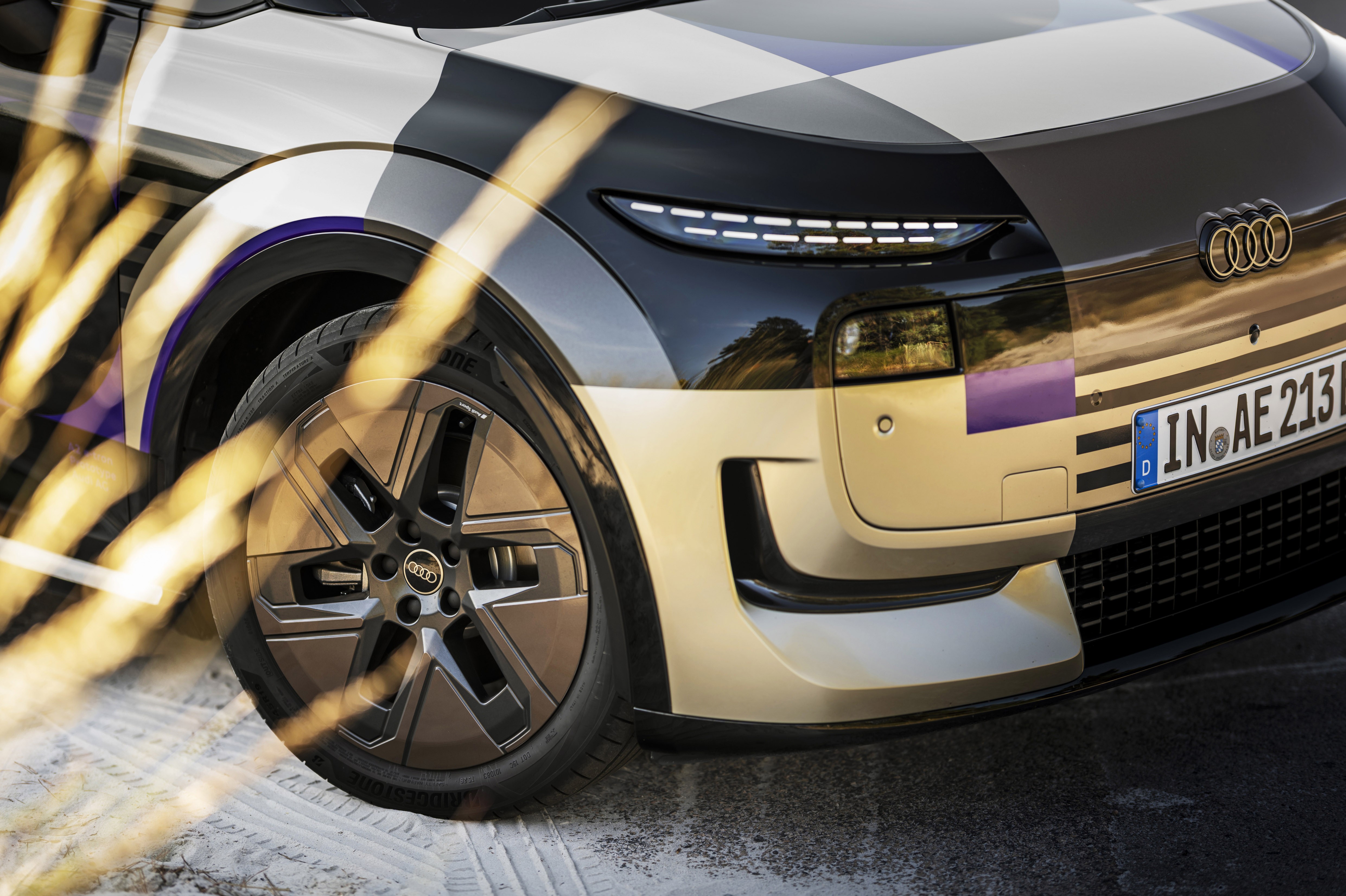 Parte frontal de um carro Audi com camuflagem quadriculada preta, branca e roxa, destacando o farol de LED, a grade e a roda com o logotipo da marca. A placa de licença alemã IN AE 213 está visível. Focos de luz dourada atravessam a imagem