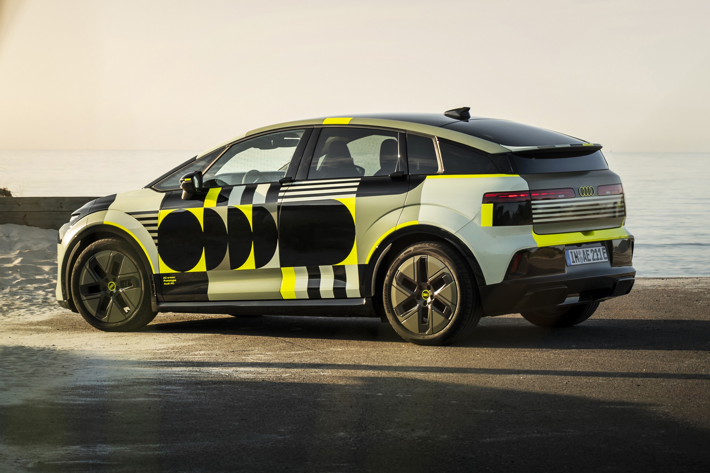 Carro elétrico camuflado com padrões geométricos em preto, amarelo e cinza claro, estacionado em uma estrada de asfalto perto do mar, sob luz solar