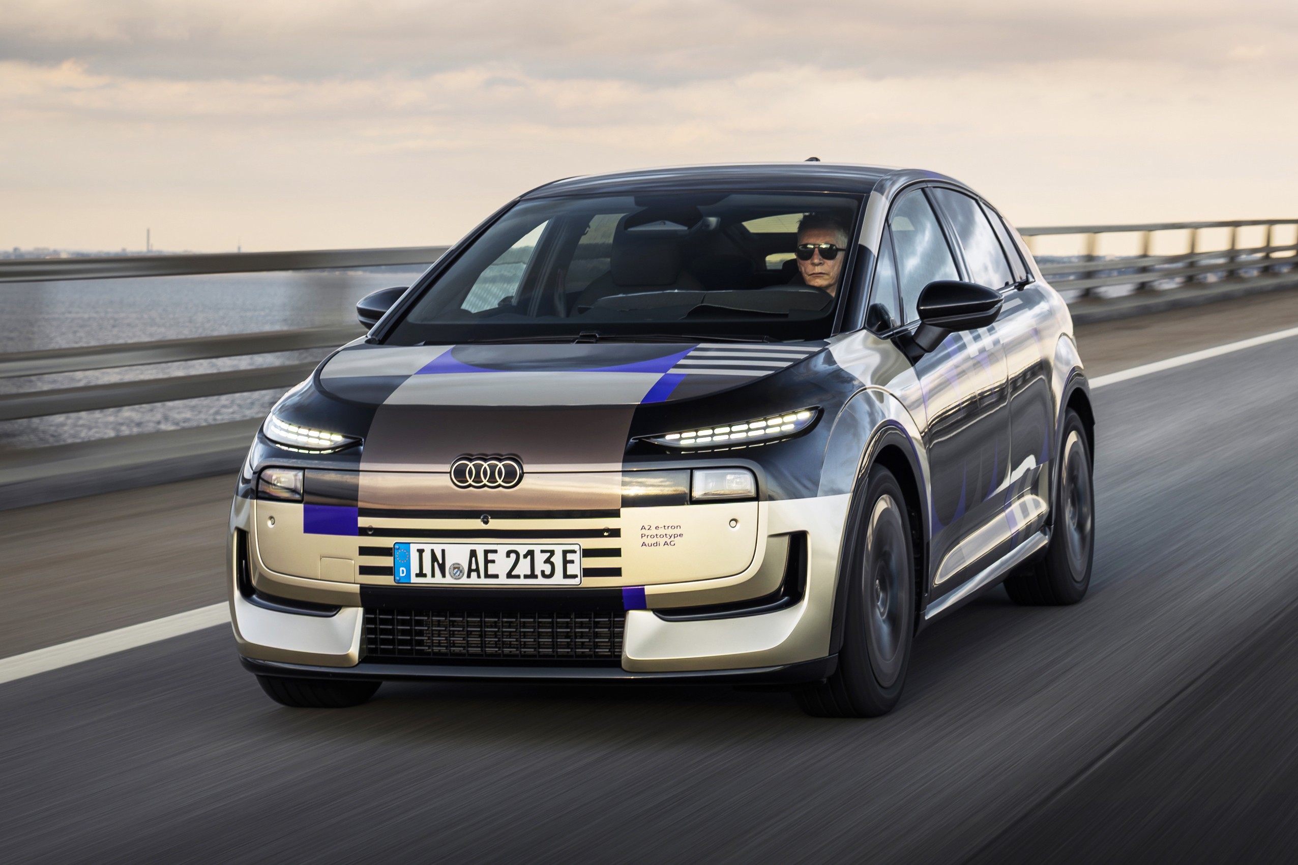 Carro Audi elétrico camuflado com padrões geométricos em tons de preto, azul e bege, dirigido por uma pessoa de óculos escuros, em movimento rápido sobre uma ponte com água ao fundo