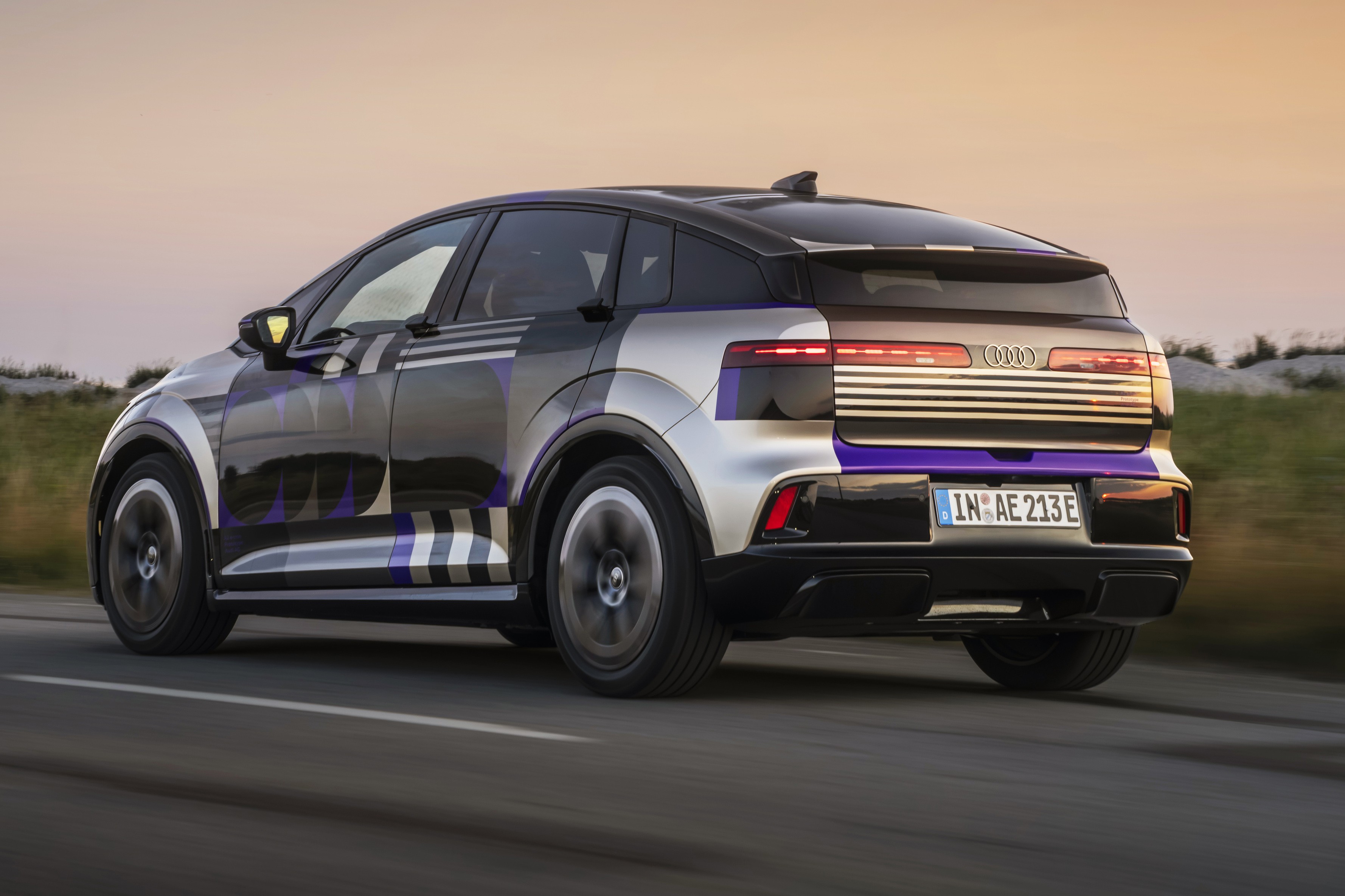 Carro Audi elétrico camuflado com padrões geométricos em preto, prata e roxo, visto de traseira lateral, em movimento numa estrada, com céu claro ao fundo