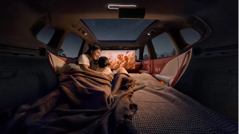 Mãe e filha assistem a um filme em um carro adaptado para acampamento, com o banco traseiro transformado em cama e um projetor exibindo a imagem na parte de trás. O teto solar mostra o céu noturno estrelado, criando uma atmosfera aconchegante e de aventura