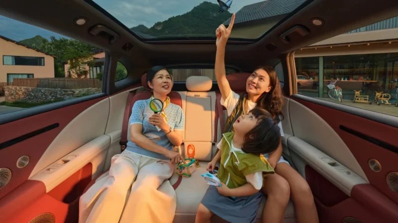 Três pessoas asiáticas, uma mulher mais velha, uma jovem e uma criança, sentadas no banco traseiro de um carro com teto solar. A jovem aponta para uma borboleta azul voando acima, enquanto a criança e a mulher mais velha olham para cima com expressões de surpresa e alegria. A mulher mais velha segura uma lupa. O interior do carro é luxuoso, com estofamento em tons de creme e bordô. Ao fundo, casas e montanhas sob um céu azul.