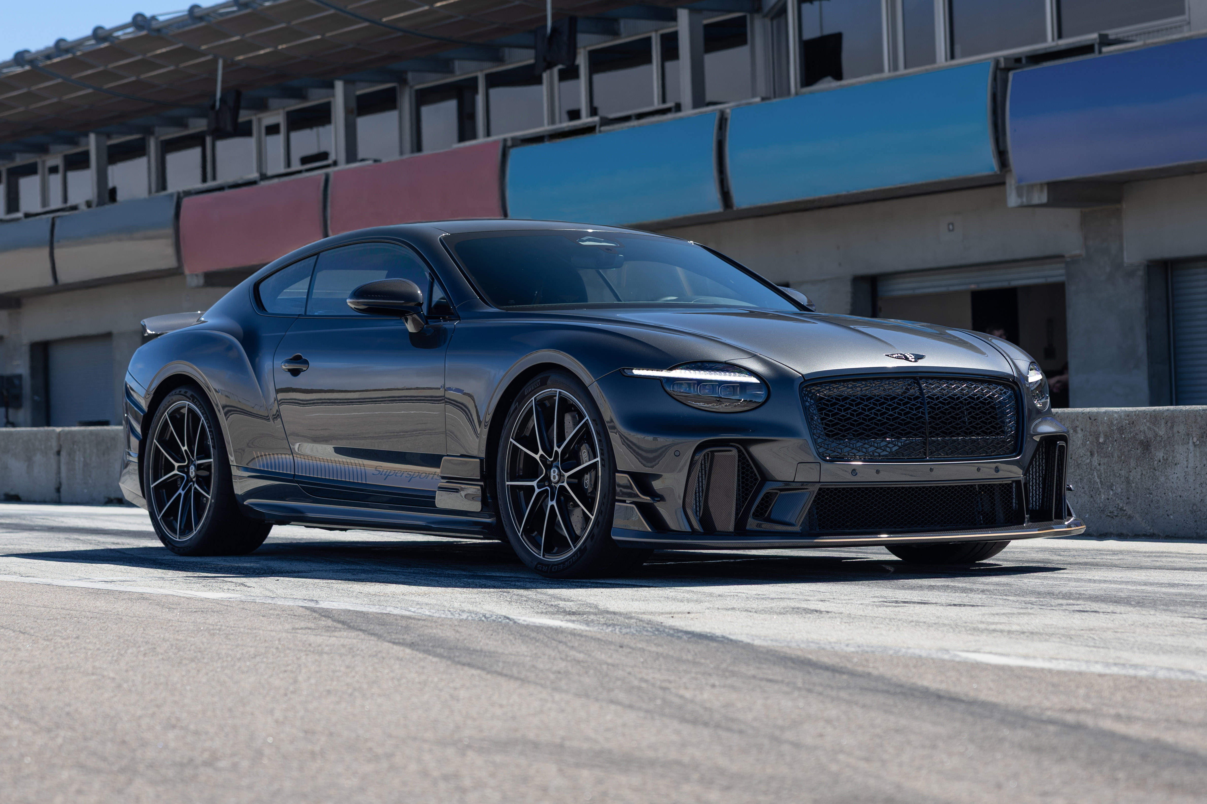 Carro esportivo Bentley Continental GT preto fosco estacionado em uma pista de corrida, com detalhes em fibra de carbono e rodas pretas. Ao fundo, uma arquibancada com painéis coloridos em azul e vermelho