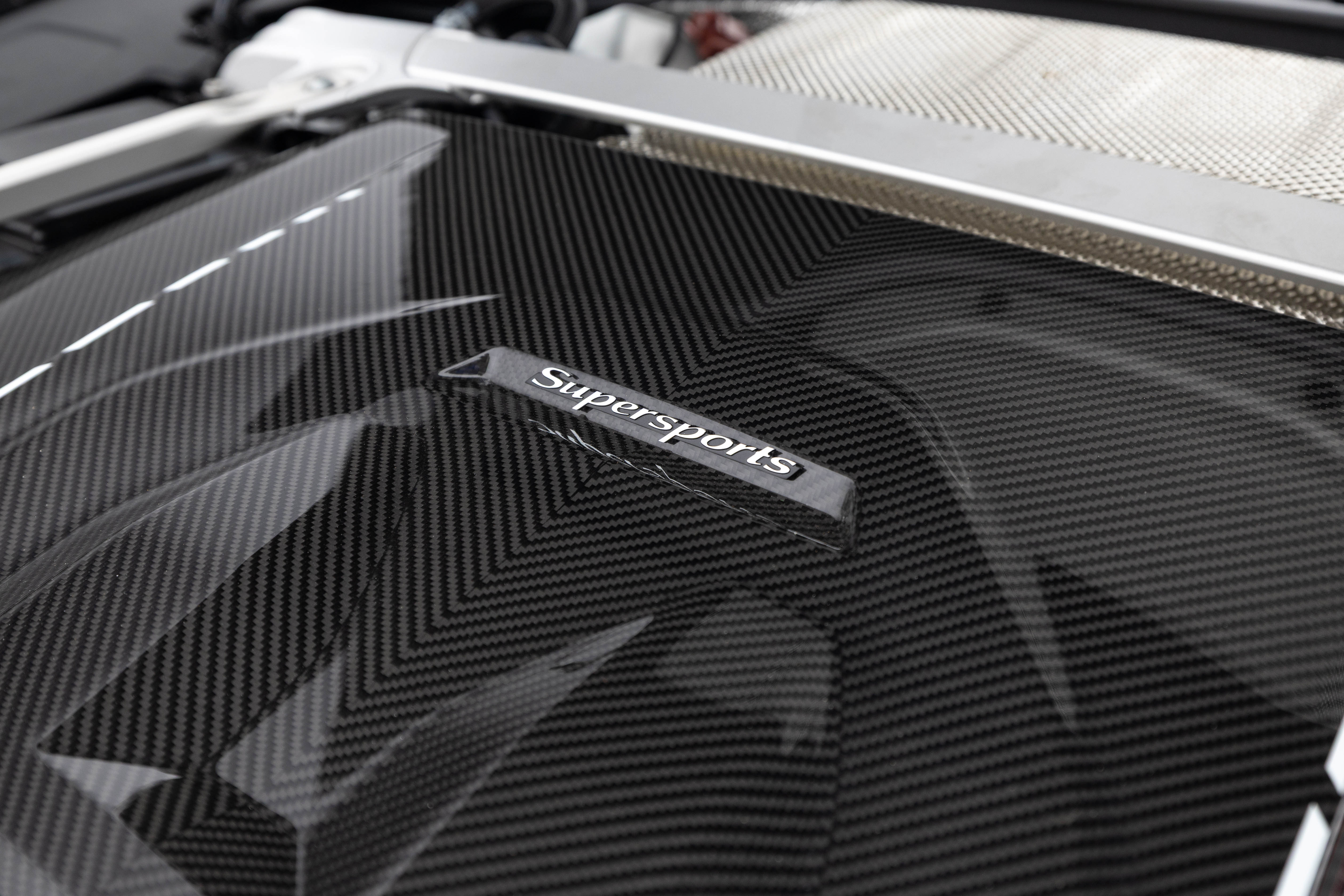 Detalhe do motor de um carro esportivo, com uma tampa preta de fibra de carbono e o emblema Supersports em relevo prateado. Ao fundo, uma grade metálica prateada.