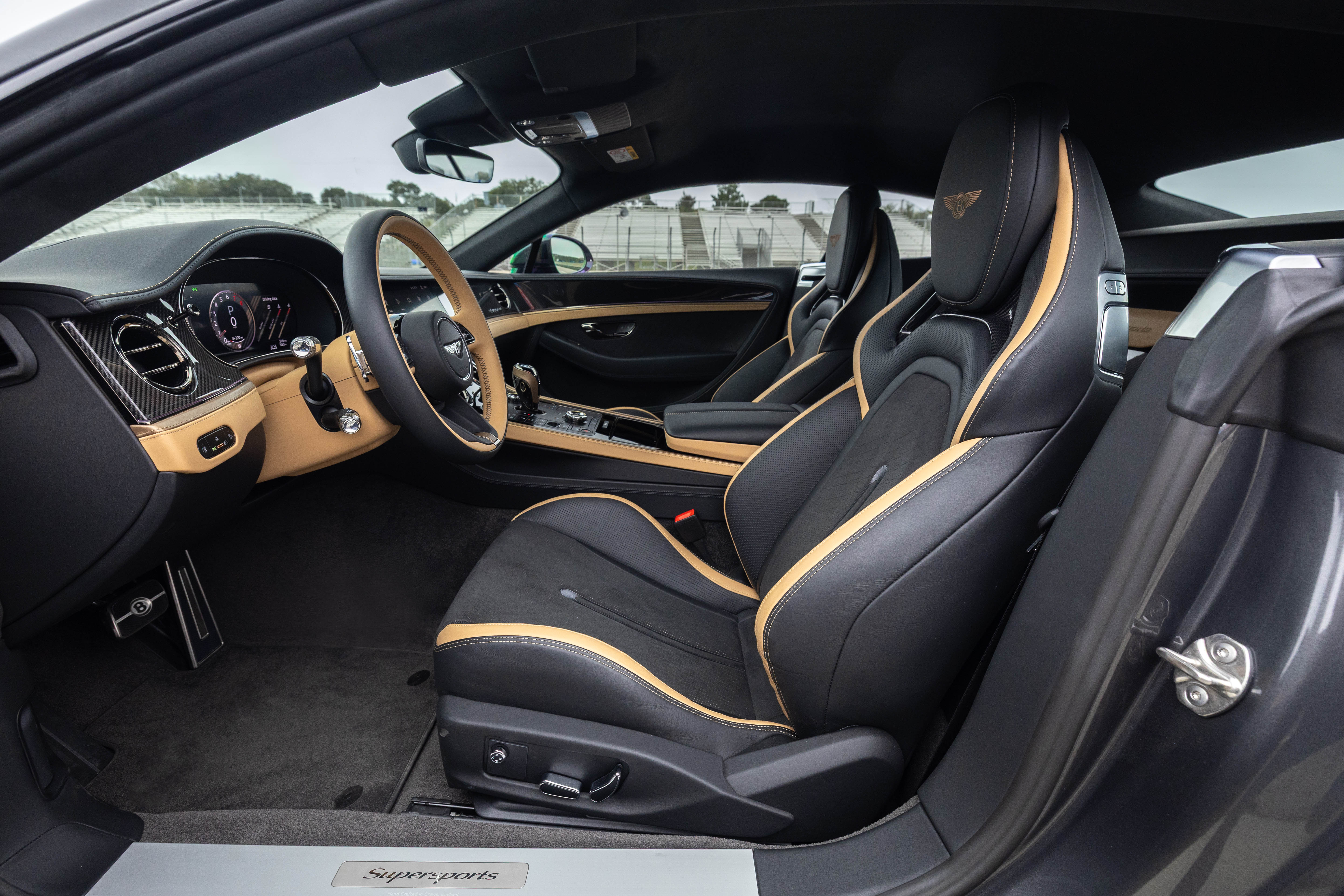 Interior de um carro de luxo com bancos esportivos pretos e detalhes em couro bege, volante bege e painel com acabamento em fibra de carbono e madeira clara, visto do lado do passageiro. O exterior é cinza escuro e o céu está nublado