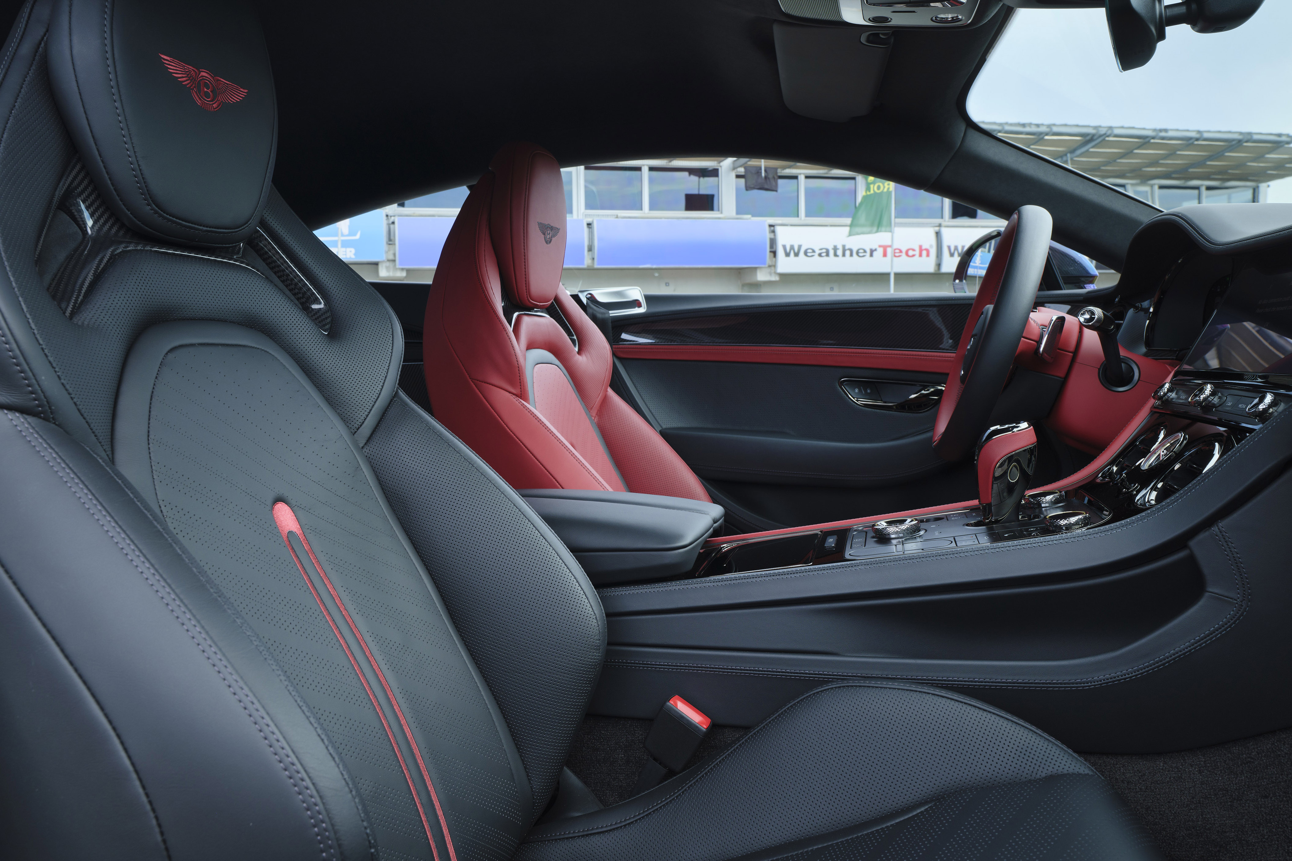 Interior de um carro esportivo de luxo, com bancos de couro preto e detalhes em vermelho. O banco do motorista, em primeiro plano, tem costuras vermelhas e um emblema de asas no encosto de cabeça. O banco do passageiro é vermelho vibrante. O painel e o volante também possuem detalhes vermelhos, com a inscrição WeatherTech visível ao fundo, através da janela