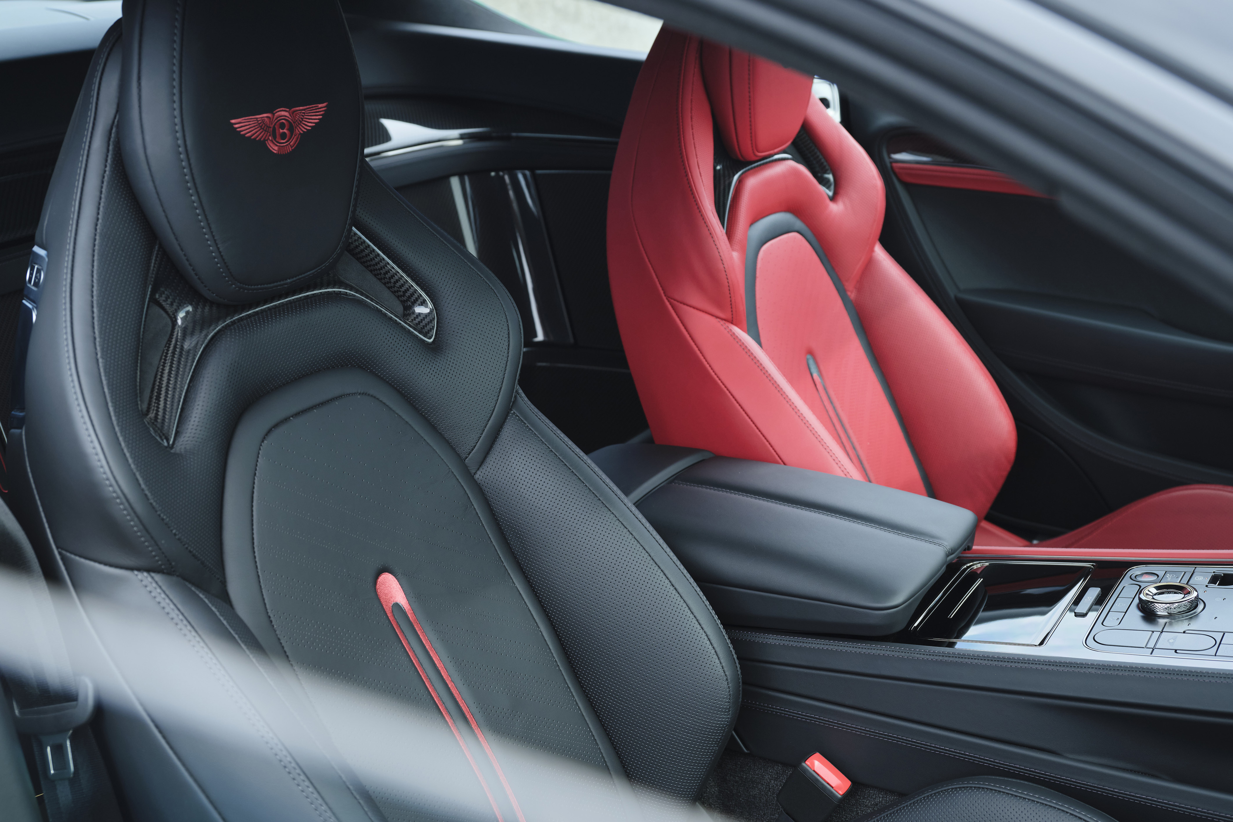 Interior de carro de luxo com bancos esportivos. O banco do motorista é preto com detalhes em fibra de carbono e o logo da Bentley em vermelho no encosto. O banco do passageiro é vermelho vibrante. Um console central preto com acabamento brilhante e detalhes cromados divide os bancos
