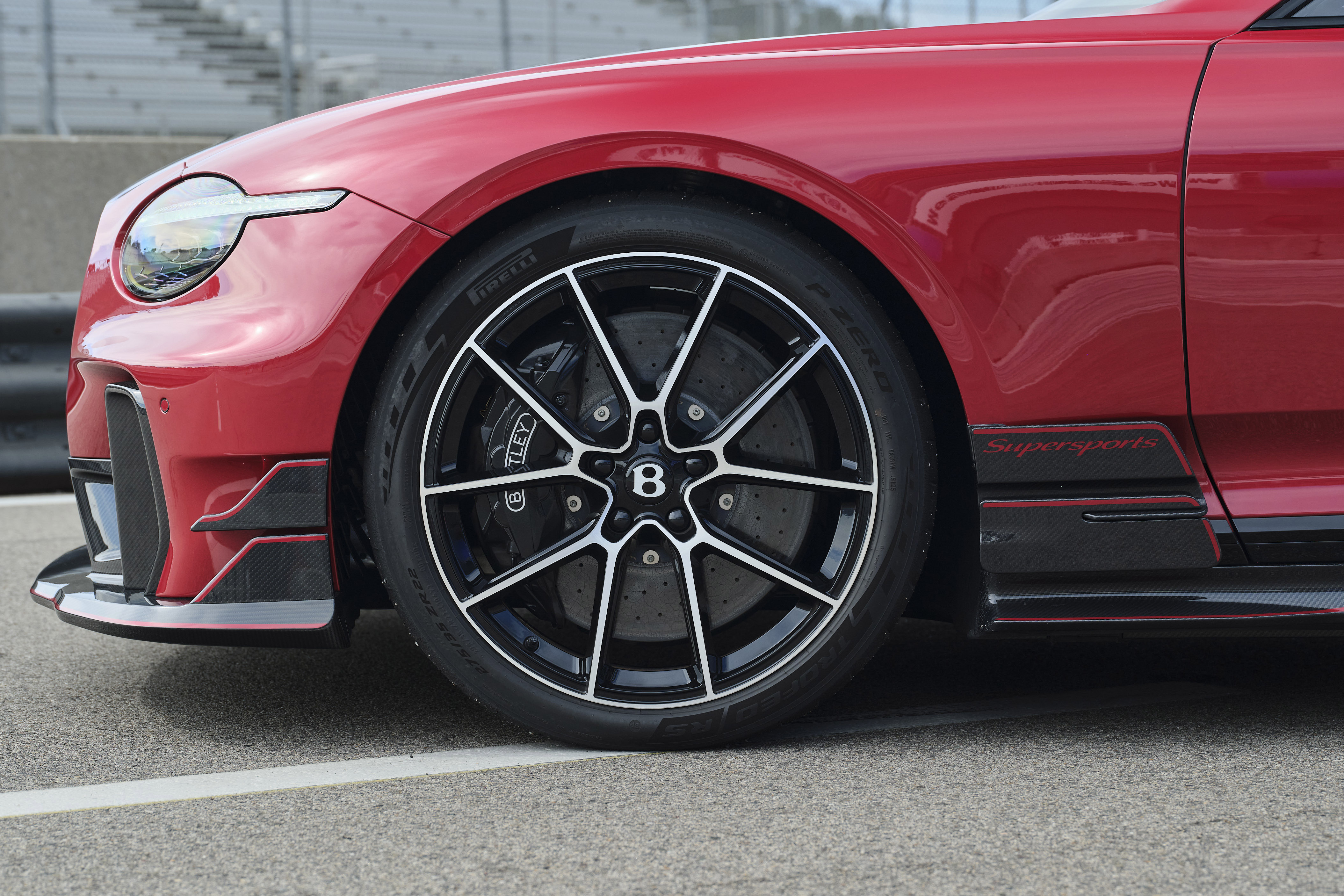 Lateral dianteira de um carro esportivo vermelho, com farol oval e detalhes aerodinâmicos pretos. A roda tem aros pretos e raios prateados, com o logo Bentley no centro. O pneu Pirelli e a inscrição Supersports em vermelho na lateral completam o visual. O veículo está em uma pista de corrida, com arquibancadas ao fundo