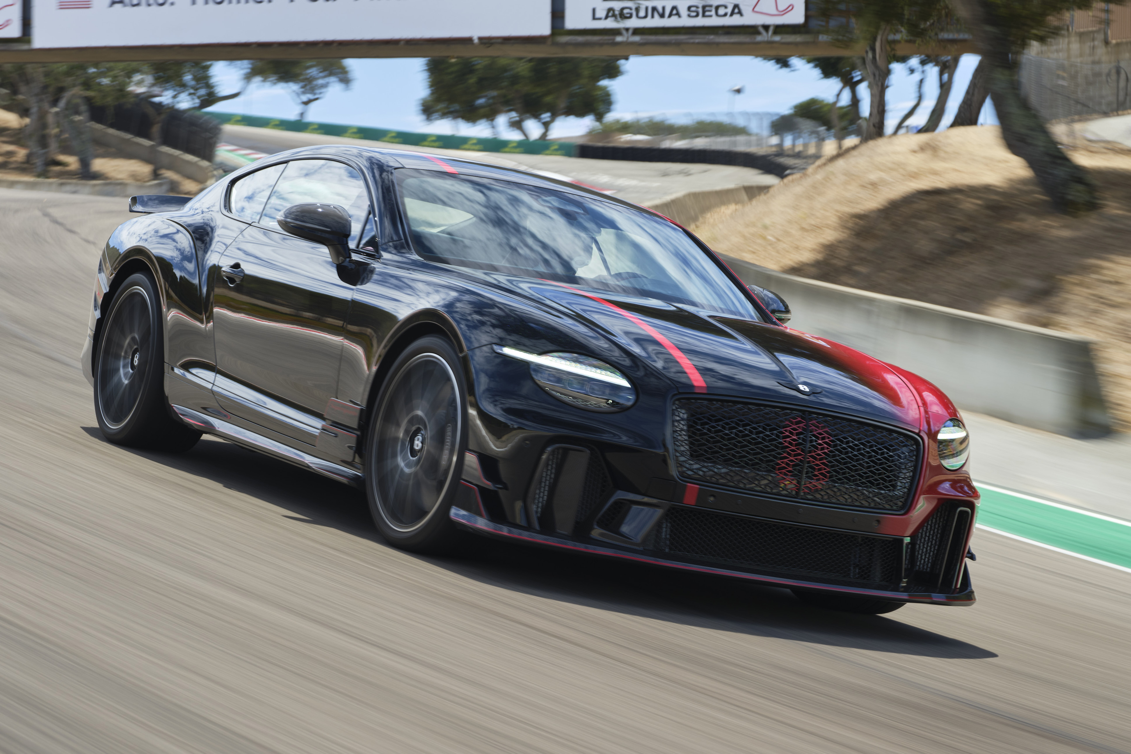 Carro esportivo Bentley preto com detalhes vermelhos e listras vermelhas no capô, em alta velocidade numa pista de corrida, com as rodas em movimento borrado. Ao fundo, árvores e um céu azul com nuvens brancas