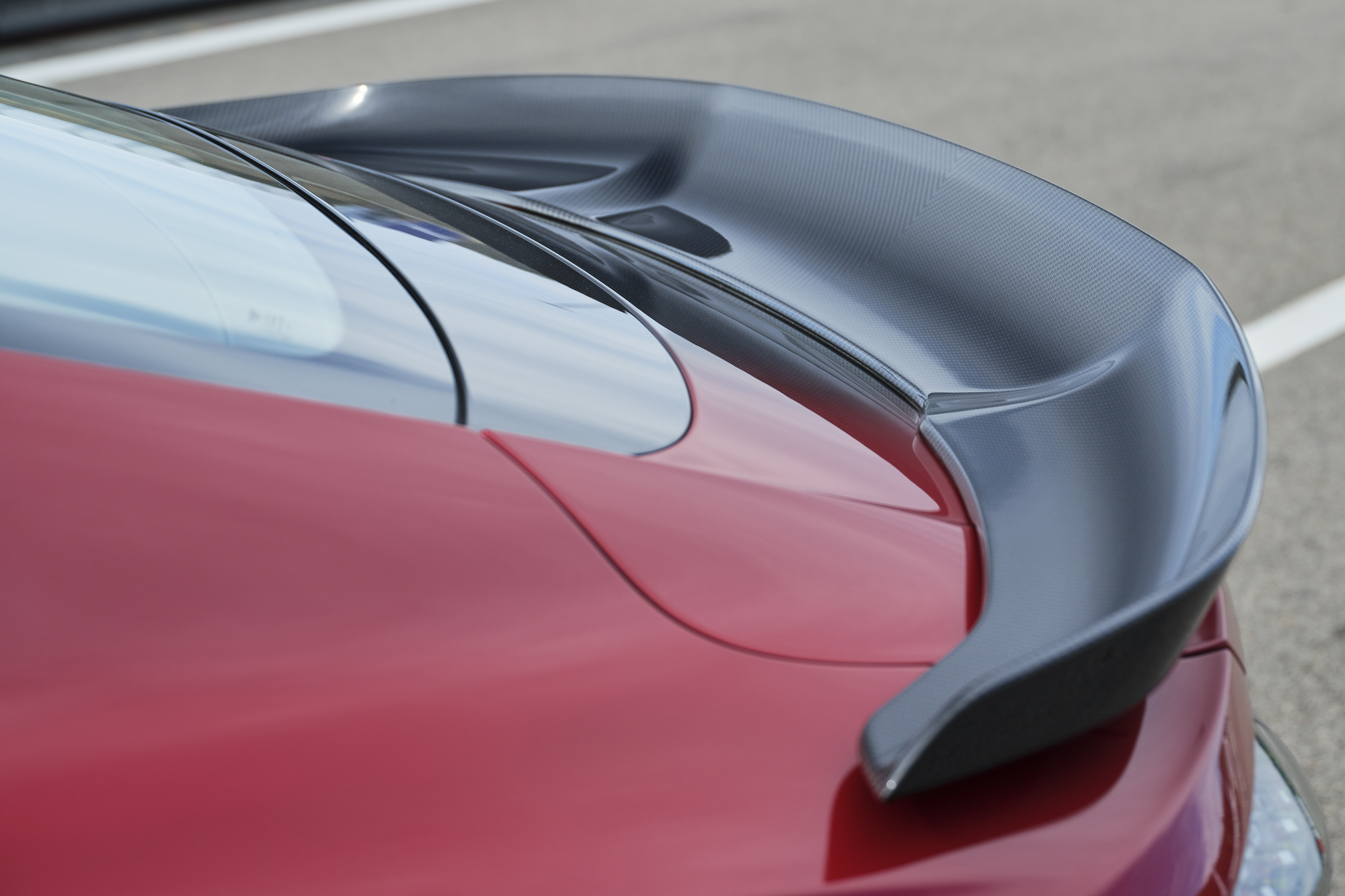 Traseira de um carro esportivo vermelho com um aerofólio preto de fibra de carbono, destacando a curvatura do design e o vidro traseiro
