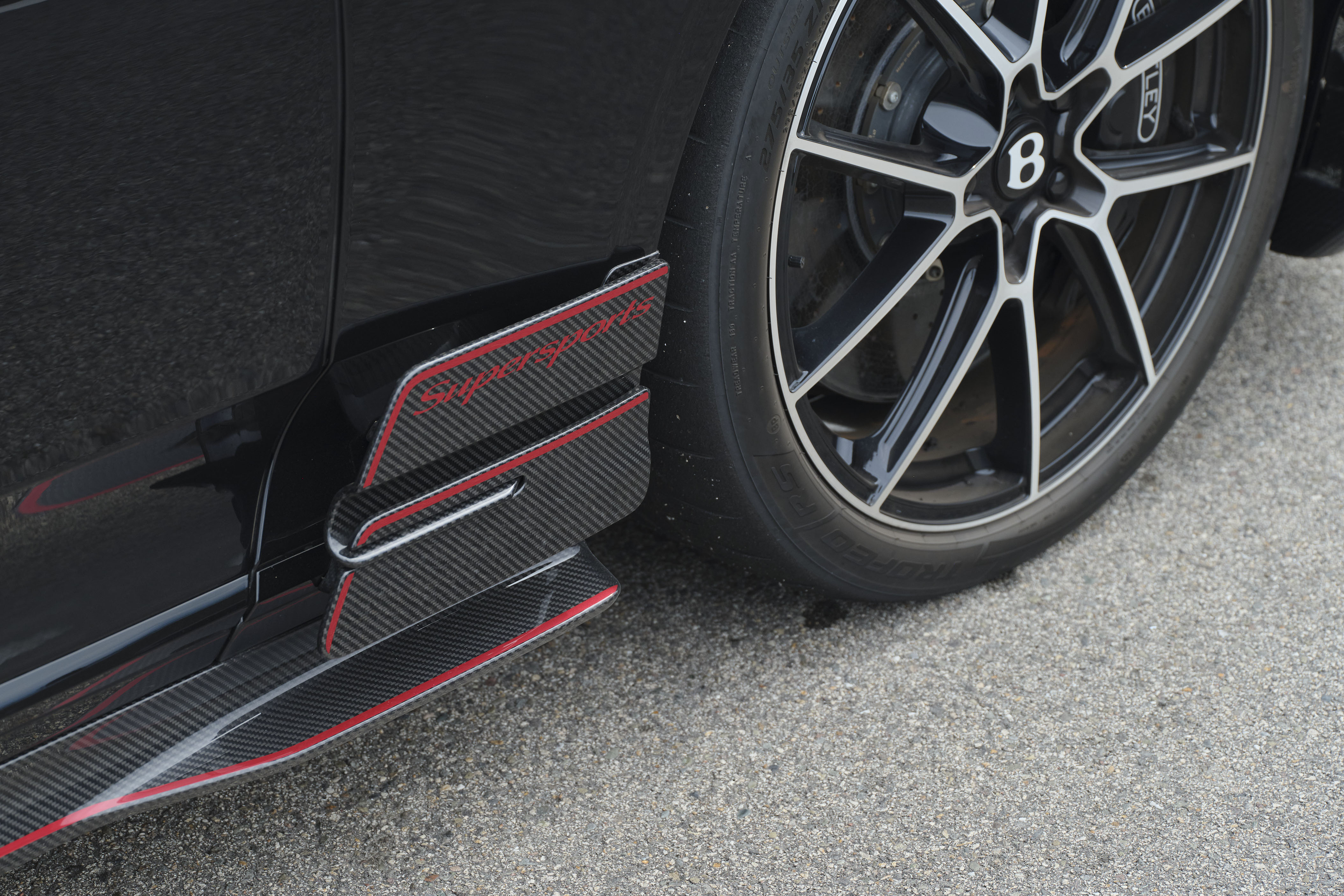 Lateral de um carro preto com detalhes em fibra de carbono e linhas vermelhas, próximo a uma roda preta com raios prateados e o logo B da Bentley no centro, sobre asfalto cinza