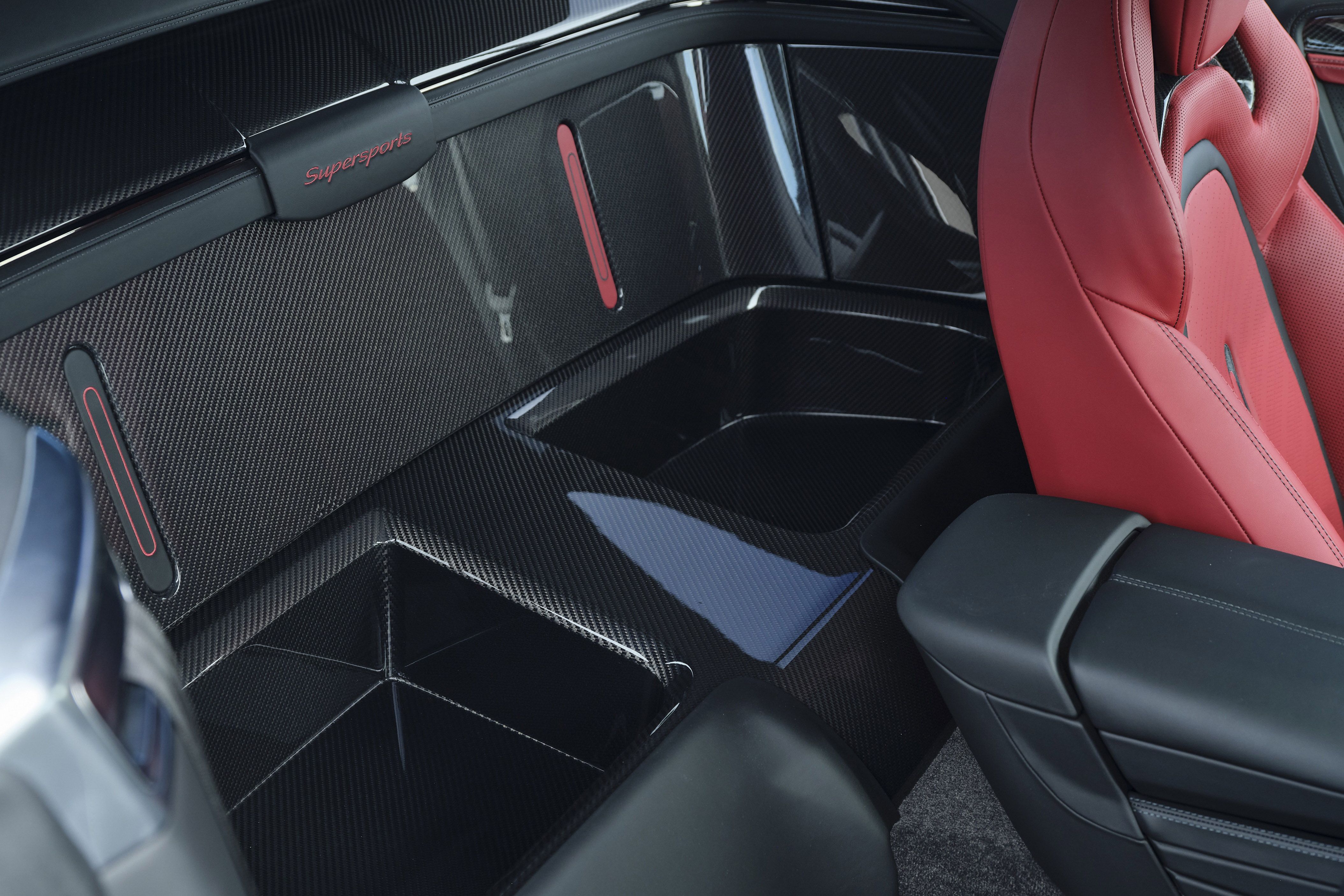 Interior de um carro esportivo com assento vermelho vibrante à direita, e painéis e compartimentos de armazenamento em fibra de carbono preta com detalhes vermelhos e azuis, exibindo a palavra Supersports
