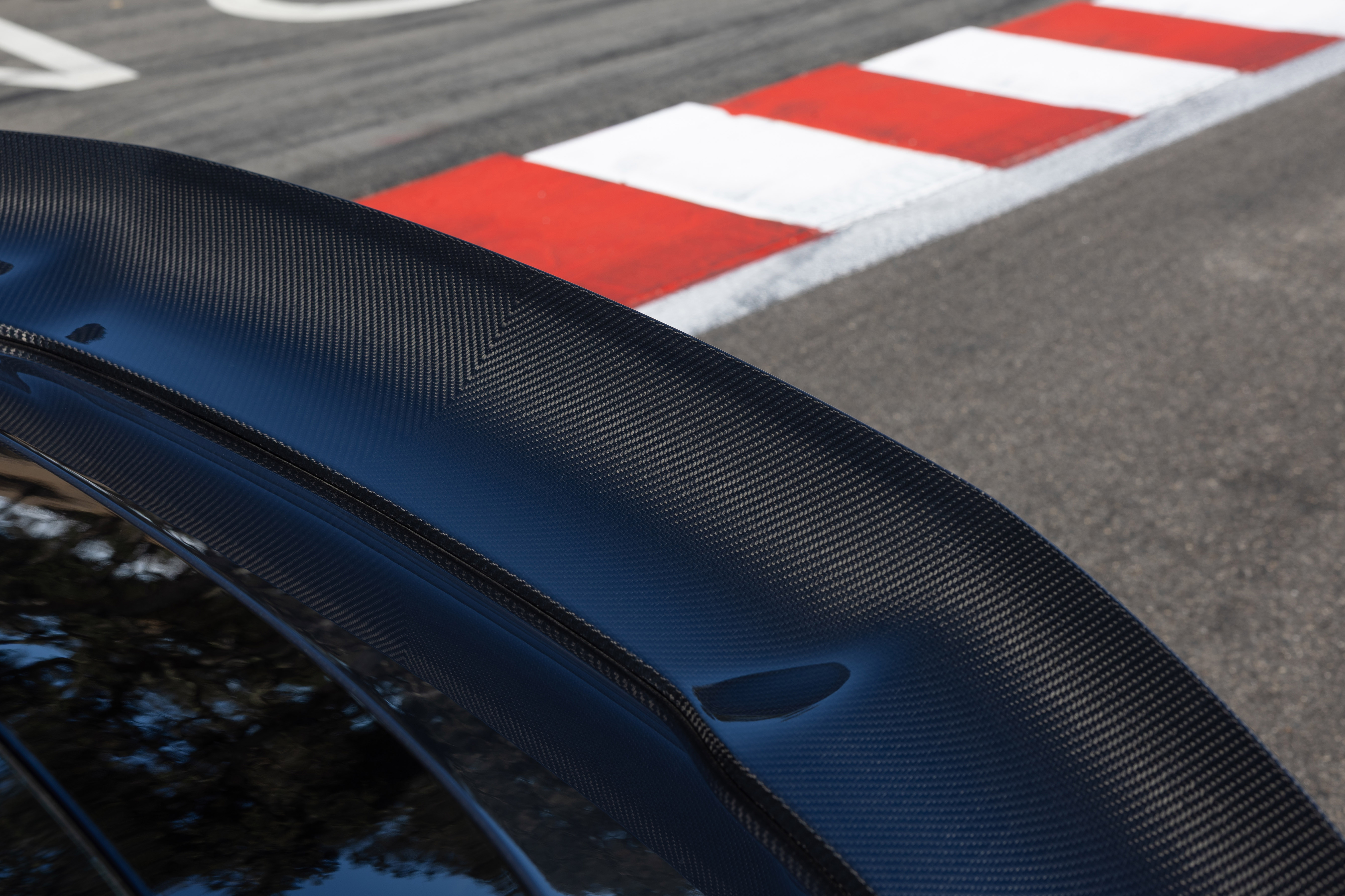 Aerofólio preto de fibra de carbono em um carro esportivo, com a textura quadriculada do material visível. Ao fundo, uma pista de corrida com zebras vermelhas e brancas