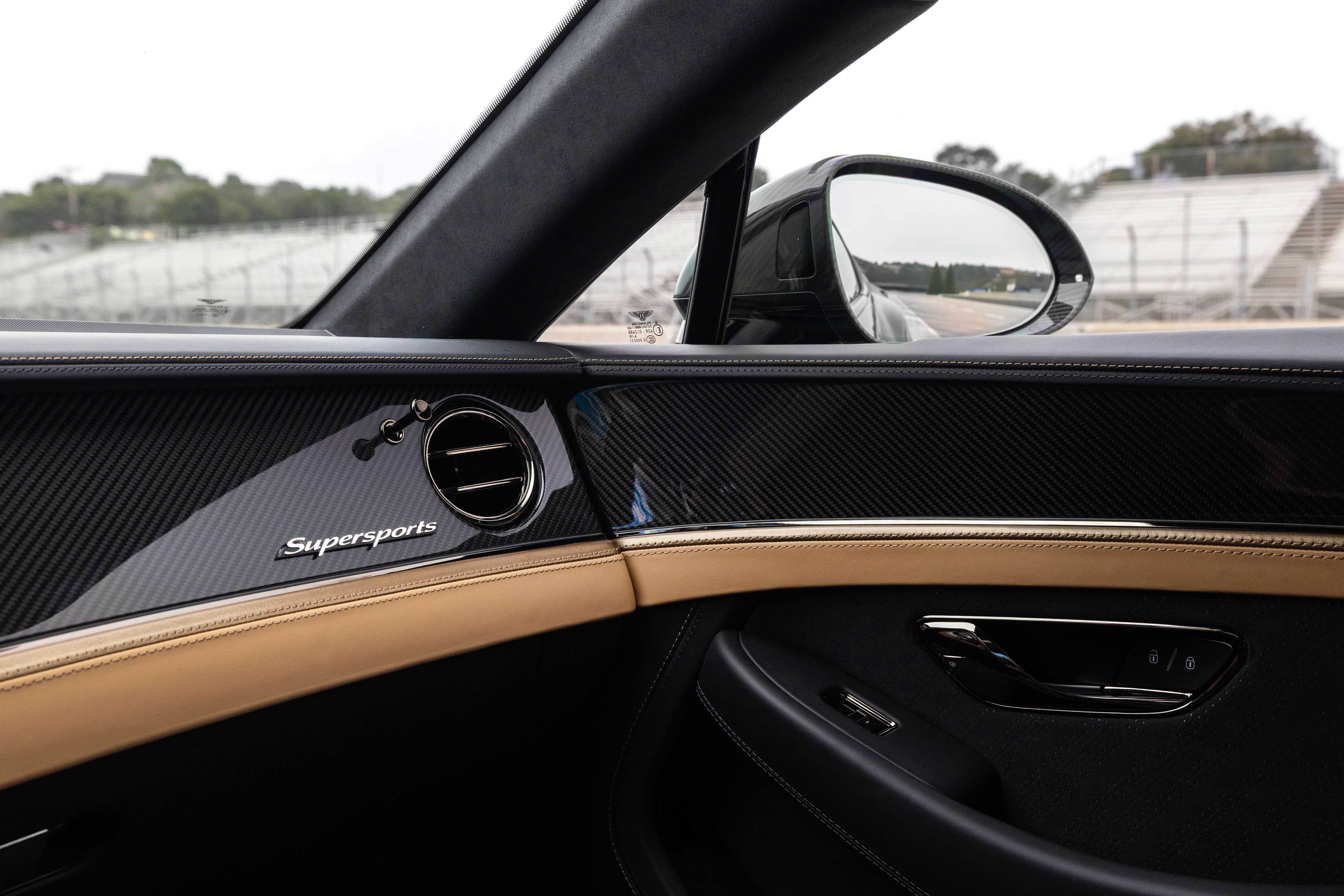 Interior de carro de luxo com painel em fibra de carbono e detalhes em couro bege, exibindo o emblema Supersports e uma saída de ar circular. Pelo retrovisor lateral, vê-se uma pista de corrida com arquibancadas ao fundo