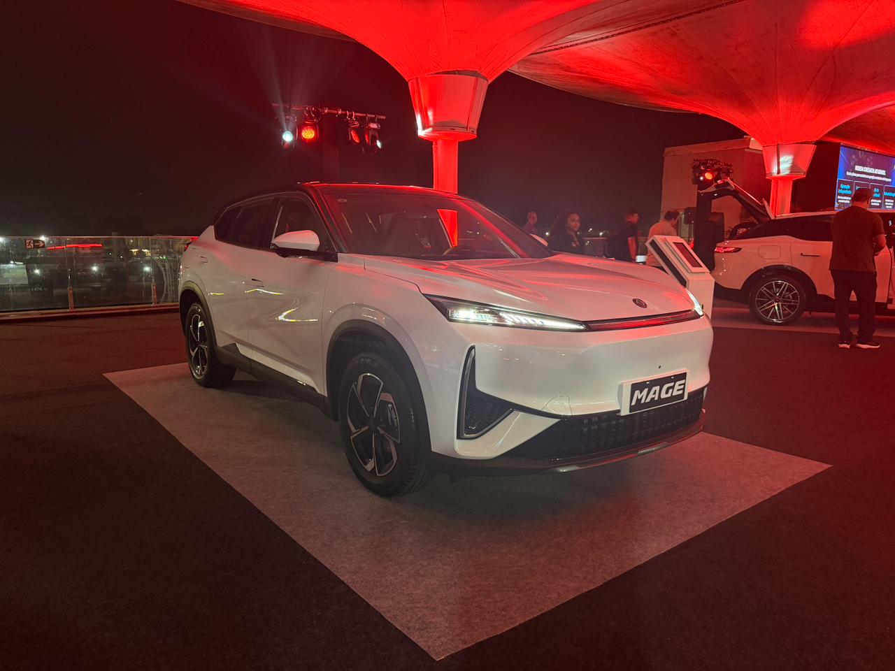 Carro SUV branco moderno, modelo Mage, em destaque sob iluminação vermelha em evento noturno. Outro carro branco e pessoas ao fundo.