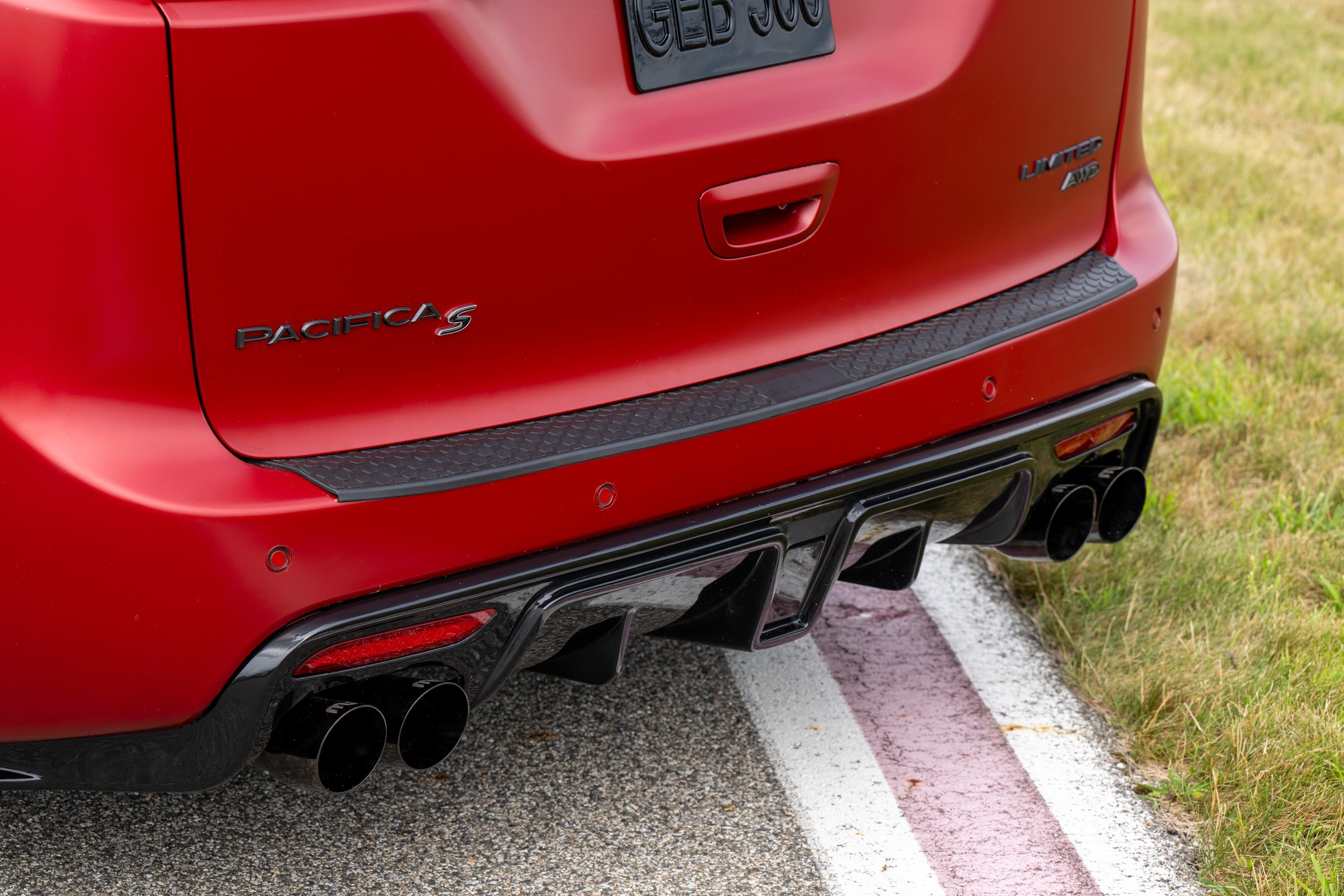Traseira de um carro vermelho com o nome PACIFICA S em relevo. Possui um para-choque preto texturizado, difusor traseiro preto com quatro saídas de escapamento e detalhes vermelhos nas luzes. O veículo está em uma pista de asfalto com faixas brancas e vermelhas, e grama verde ao lado