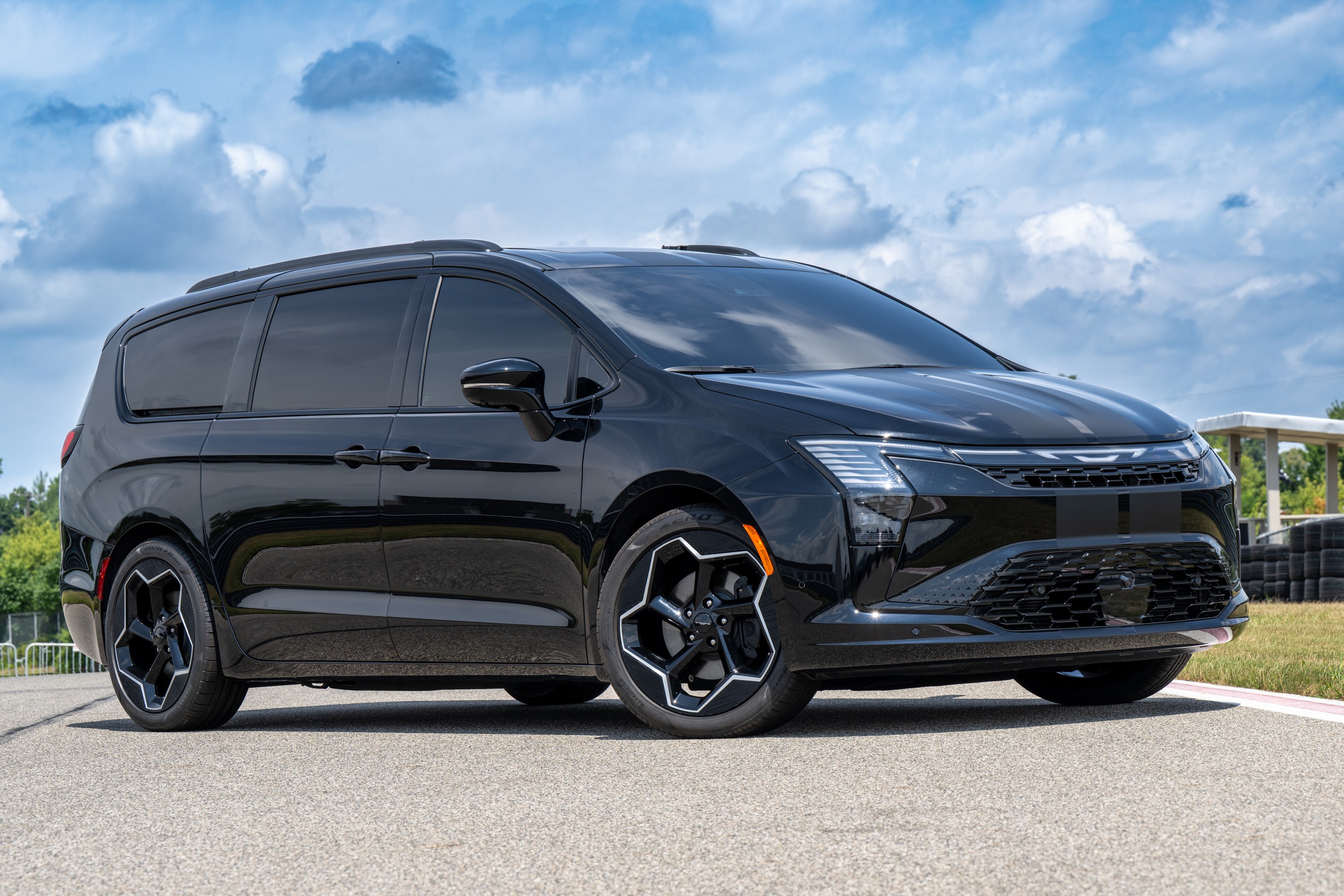 Minivan Chrysler Pacifica preta com vidros escuros e rodas esportivas pretas e brancas, estacionada em uma pista de asfalto sob céu azul com nuvens esparsas